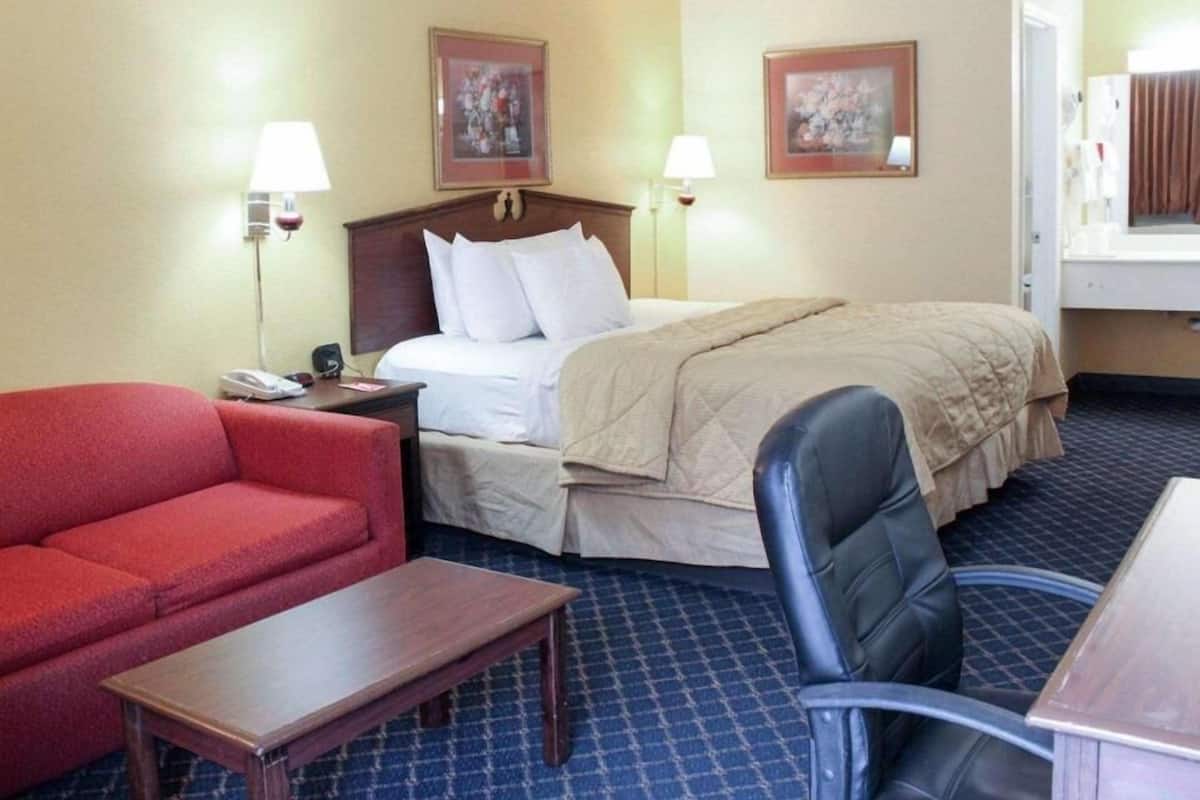 A hotel room with a bed, a red sofa, a wooden table, a black chair, and two wall-mounted lamps.