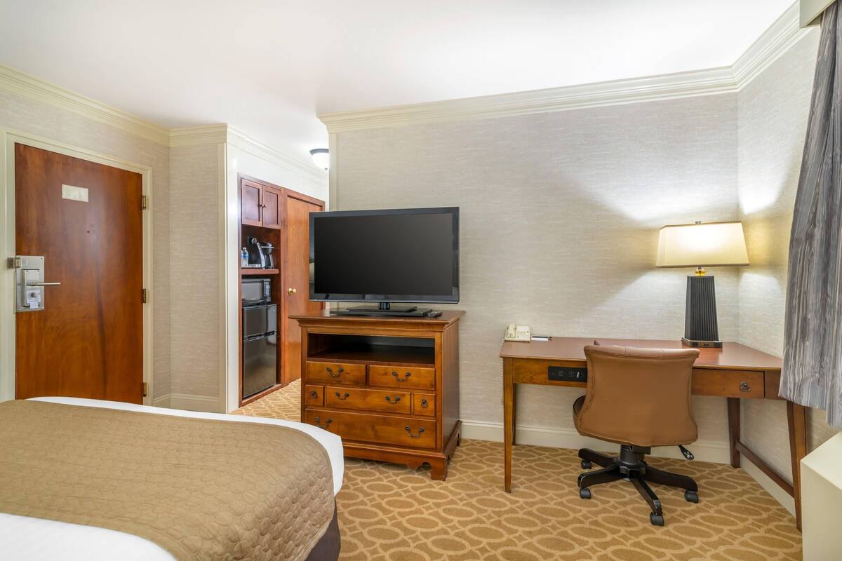 A hotel room with a bed, a desk with a chair, a television, a lamp, and a wardrobe.