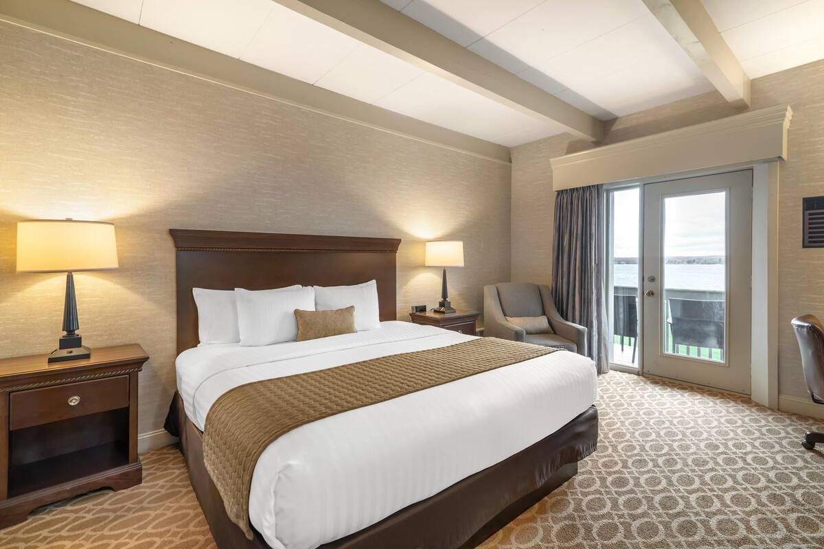 A hotel room with a large bed, two bedside tables with lamps, a chair, and a view through a sliding door.