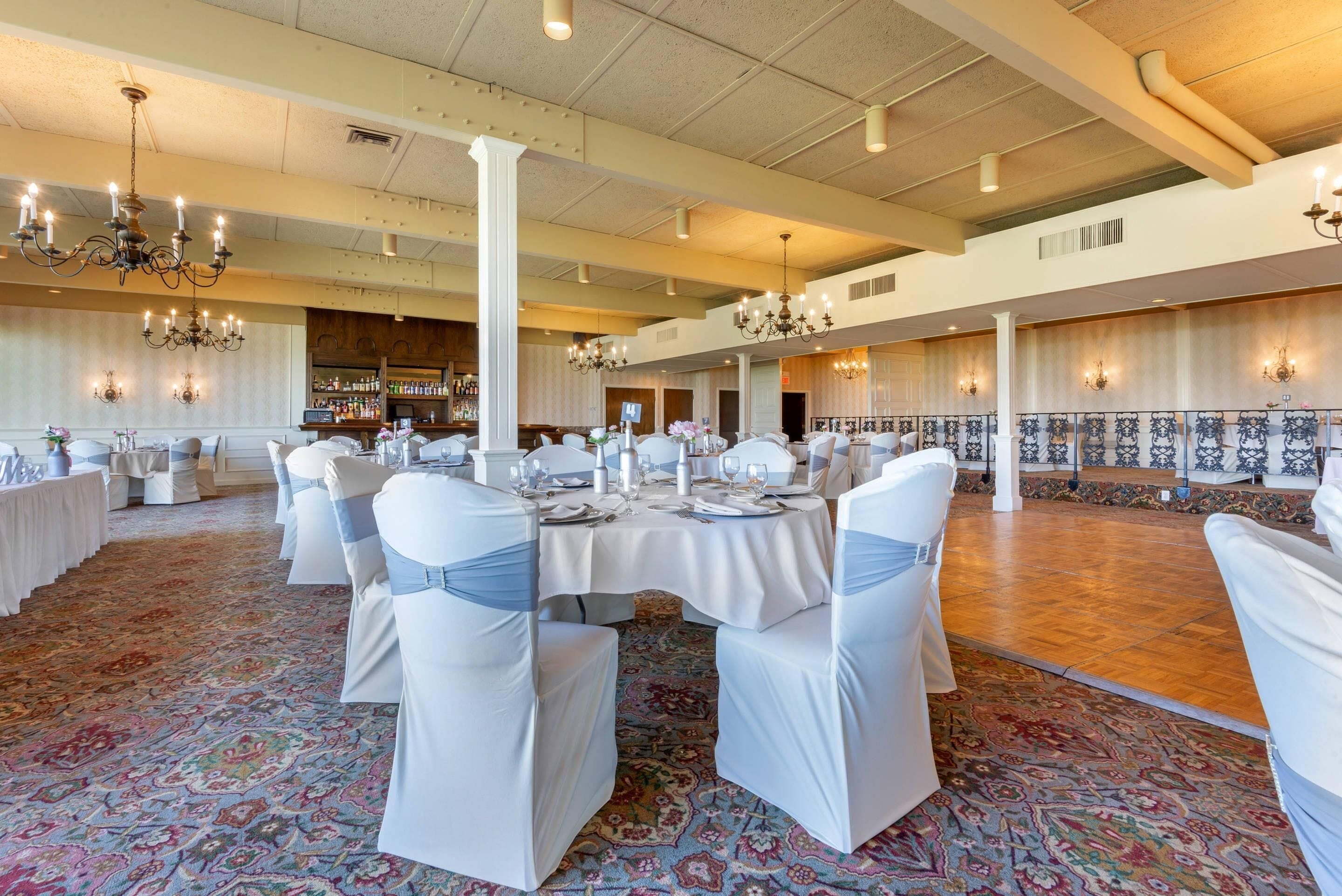 A banquet hall with round tables, white chairs, chandeliers, and a patterned carpet.