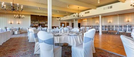A banquet hall with round tables, white chairs, chandeliers, and a patterned carpet.