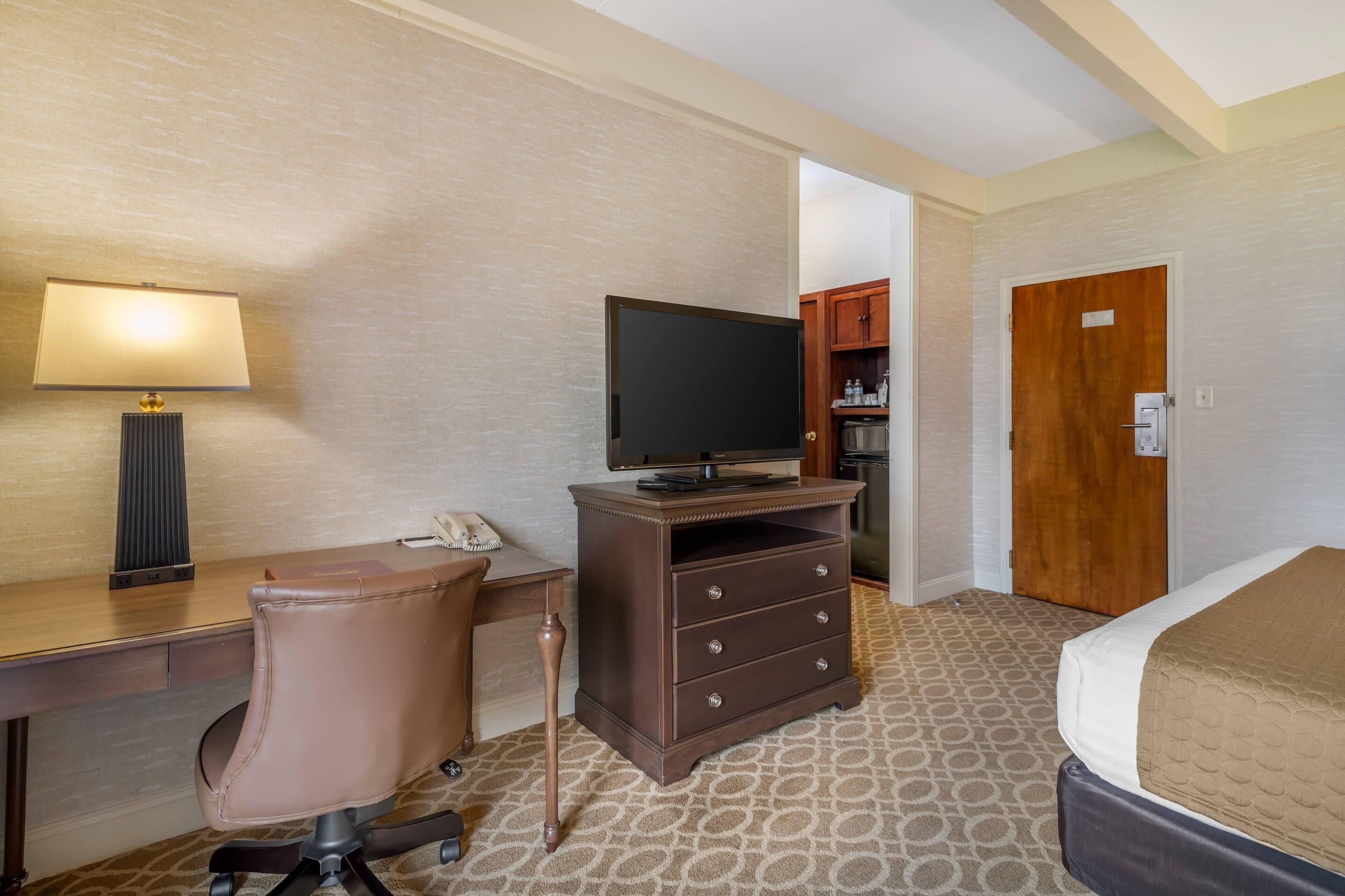 A hotel room with a desk, chair, television, dresser, and a bed.