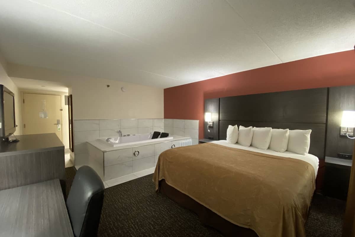 A hotel room with a bed, a desk, a chair, a bathroom with a bathtub, and a nightstand with a lamp.