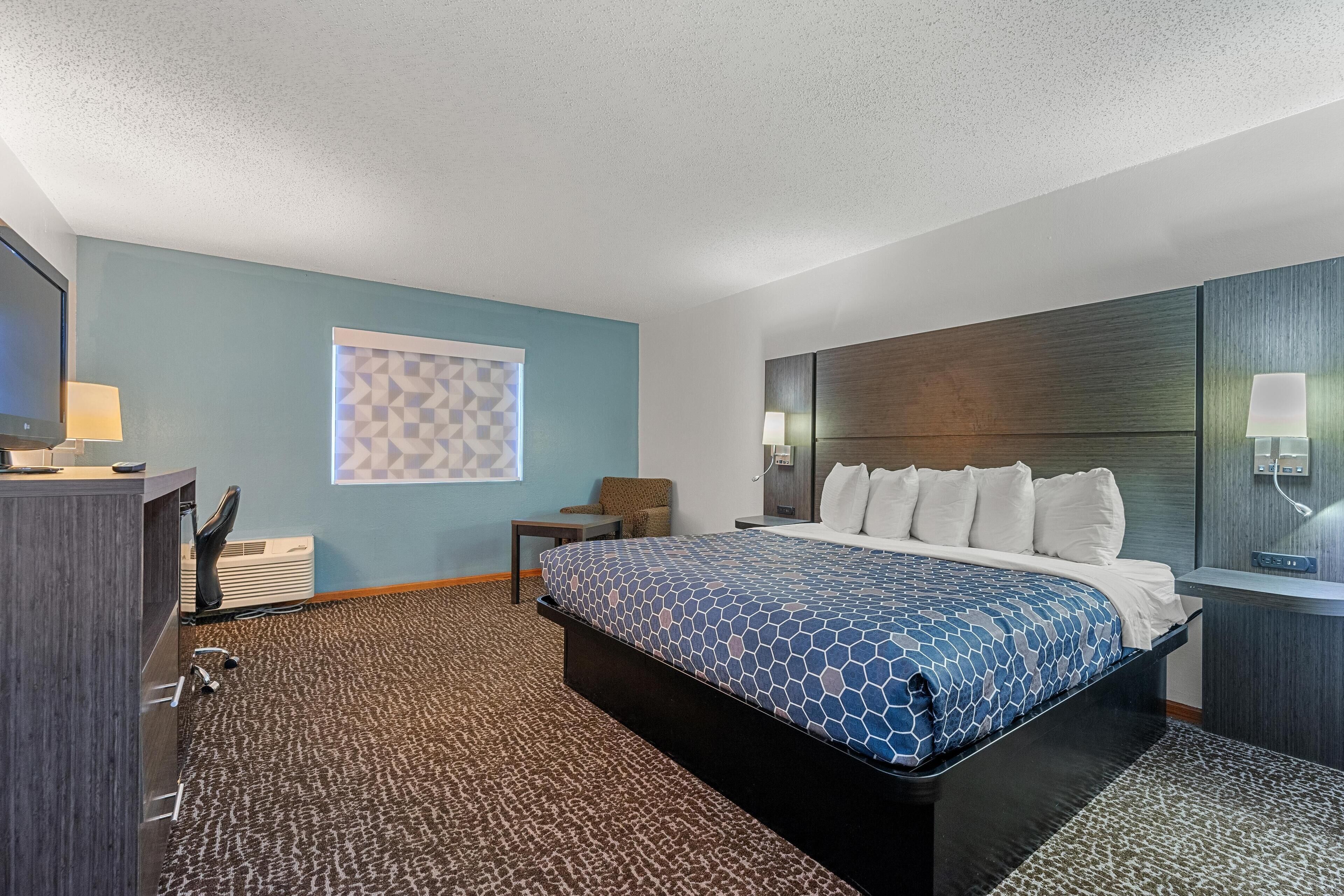 A hotel room with a large bed, a desk with a lamp, a TV, and a wall-mounted light.