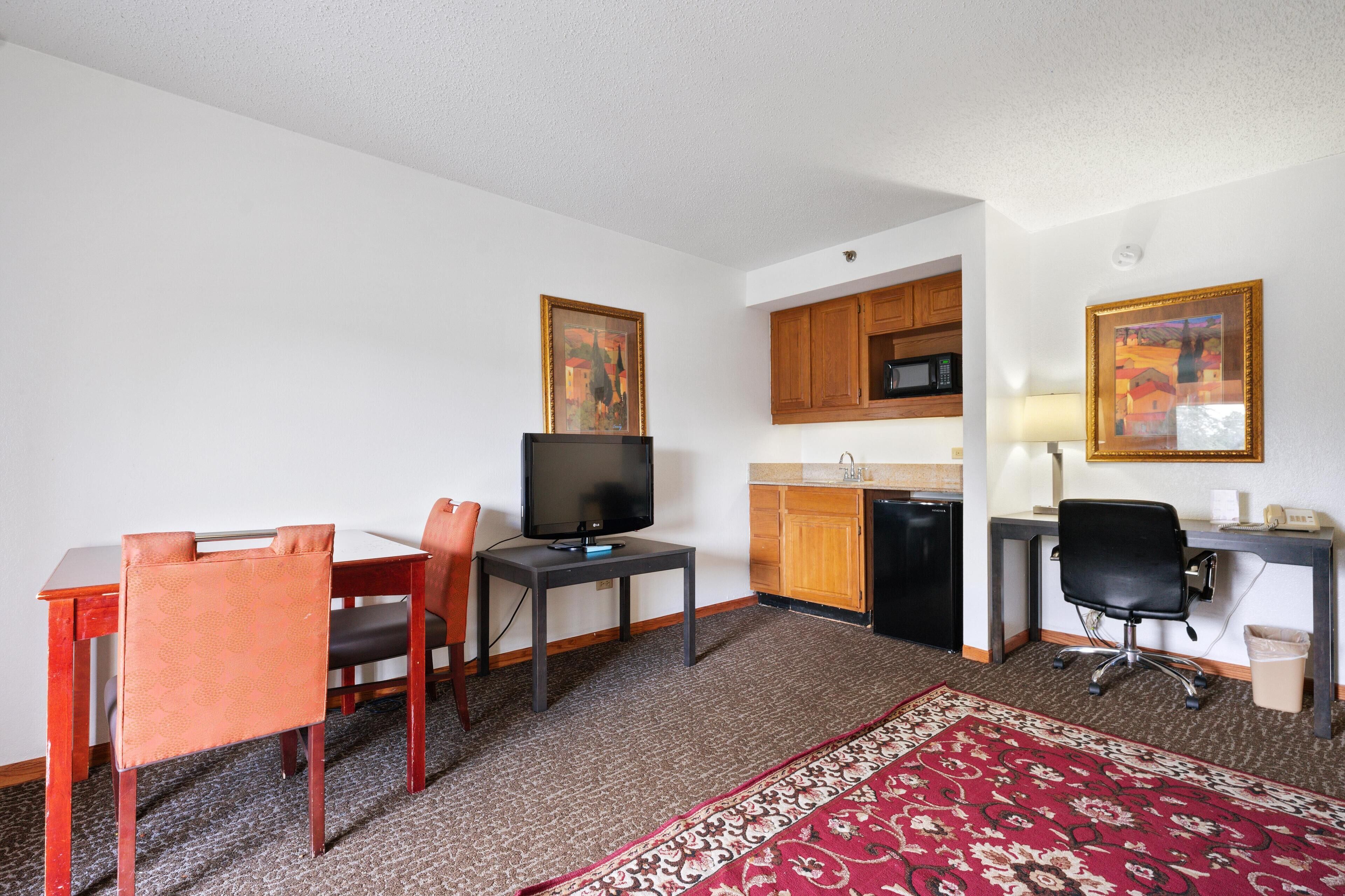 A compact hotel room with a dining table, chairs, a TV, a kitchen area with a microwave, and a desk with a chair.