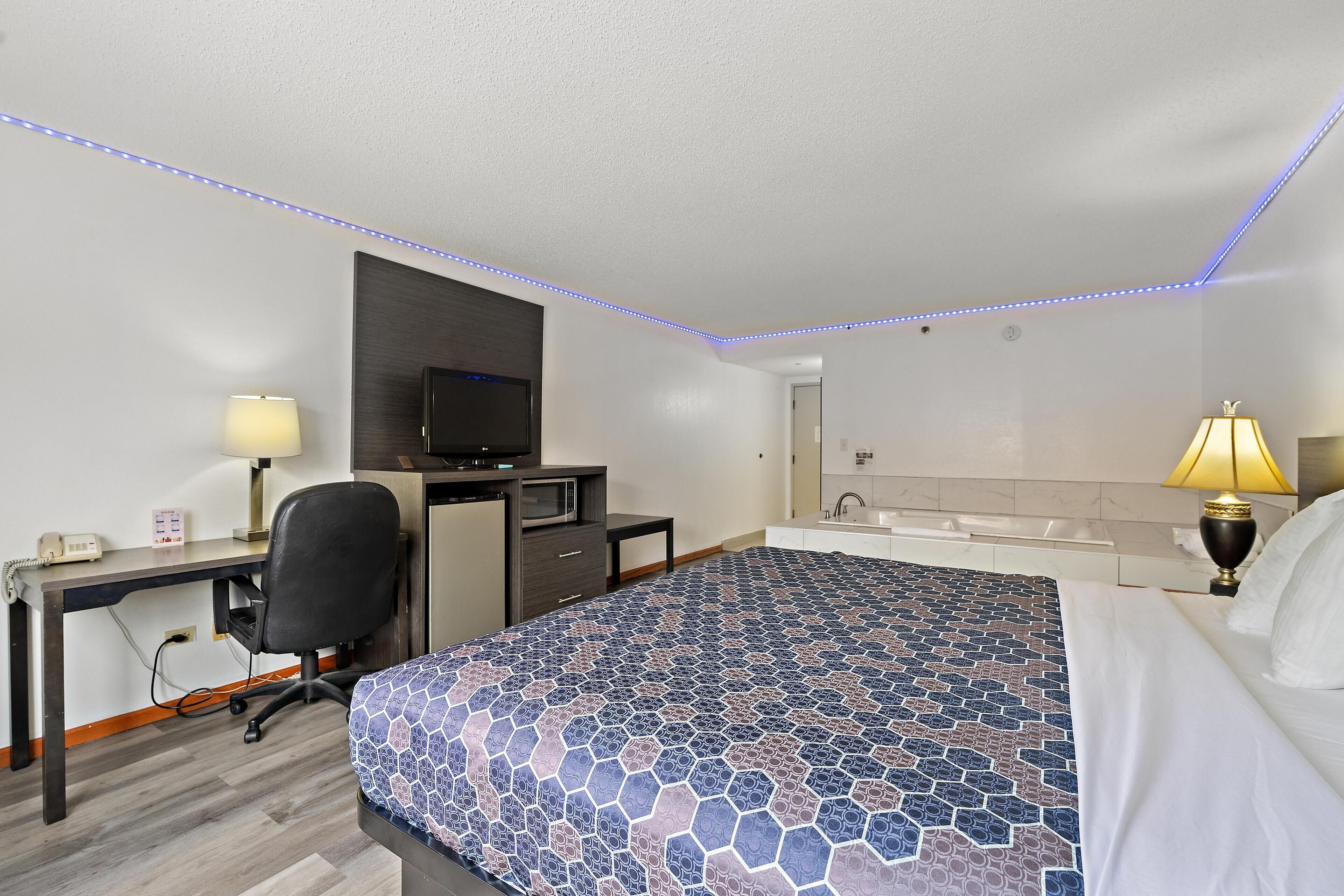 A hotel room with a bed, desk, chair, television, and a bathroom with a sink and mirror.
