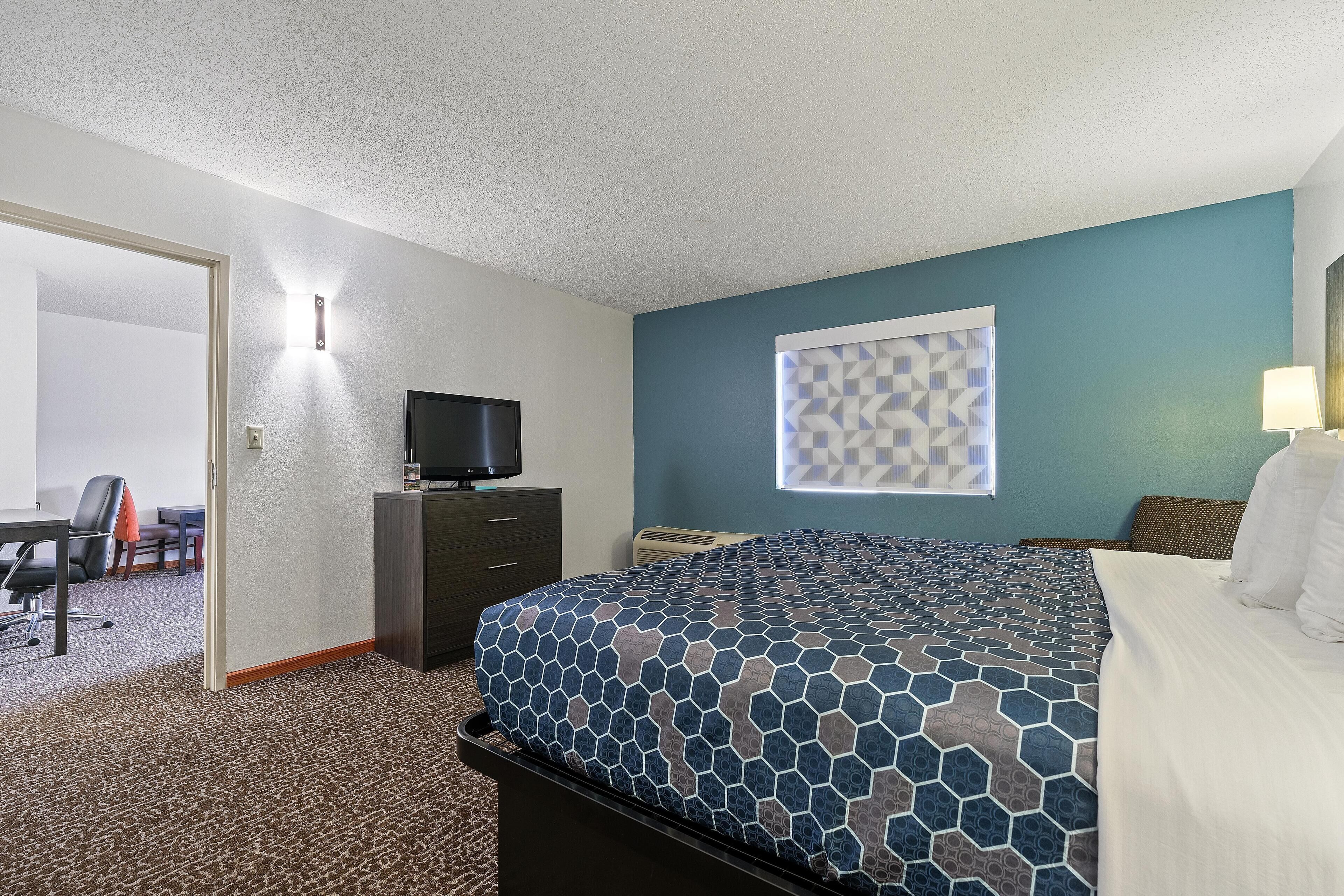 Quarto de hotel com cama, escrivaninha, cadeira, televisão e janela com padrão geométrico.