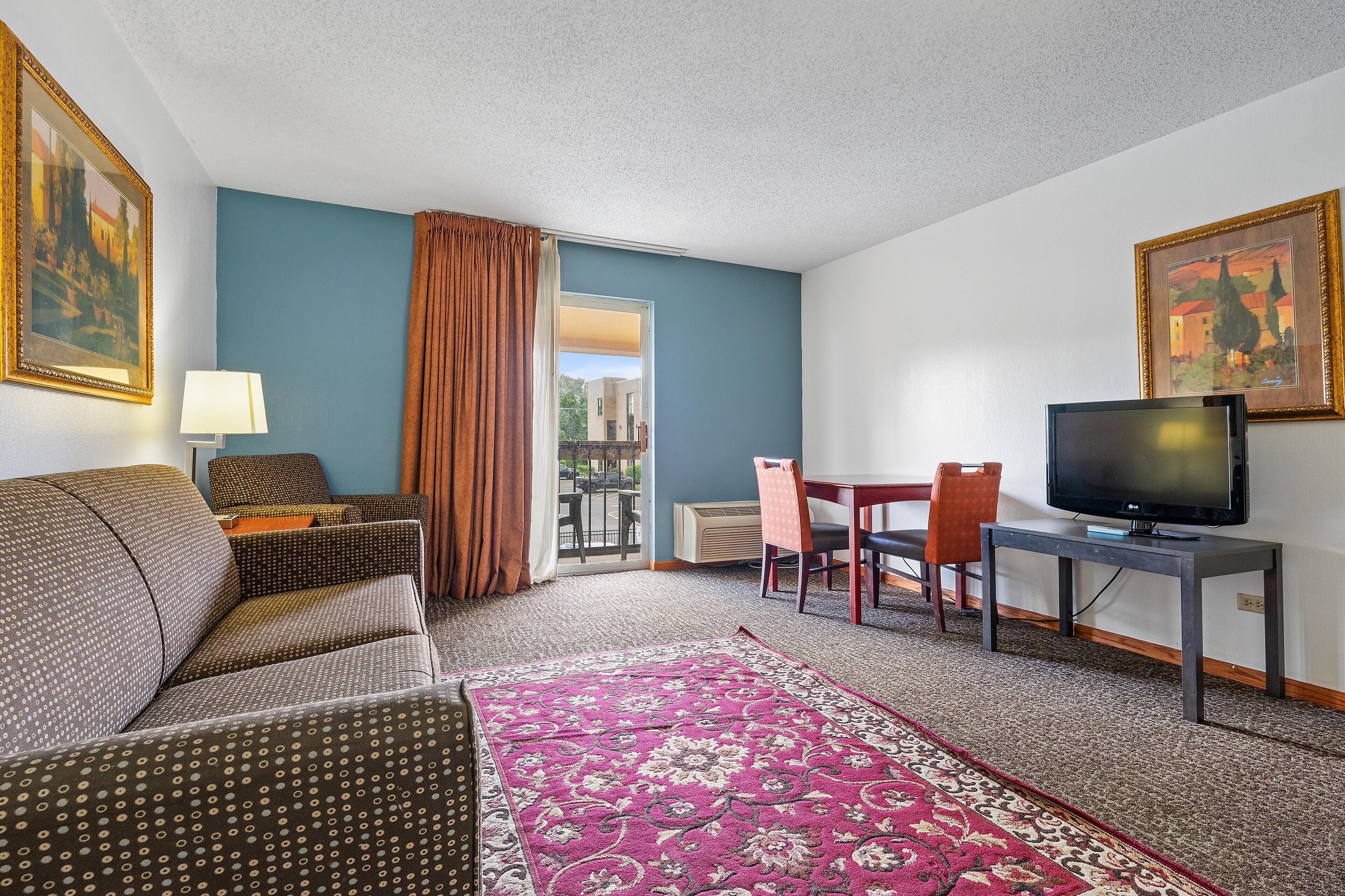 A hotel room with a sofa, a dining table with chairs, a television, and a large window with curtains.