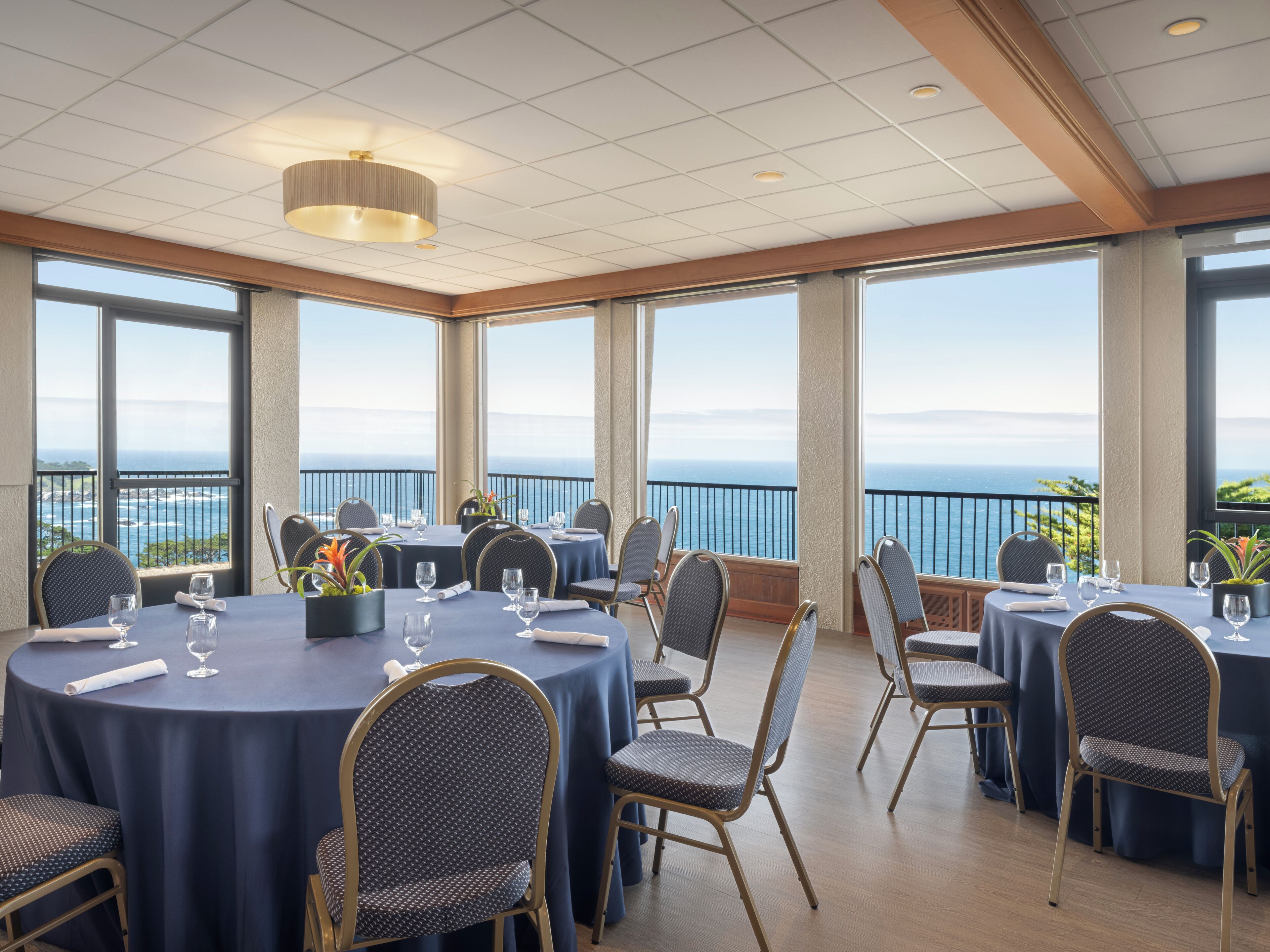 A dining area with round tables and chairs, large windows with a view of the sea, and a ceiling with recessed lighting.