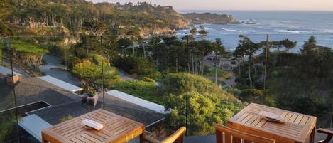 A terrace with wooden tables and chairs, overlooking a coastal landscape with trees and a view of the ocean.