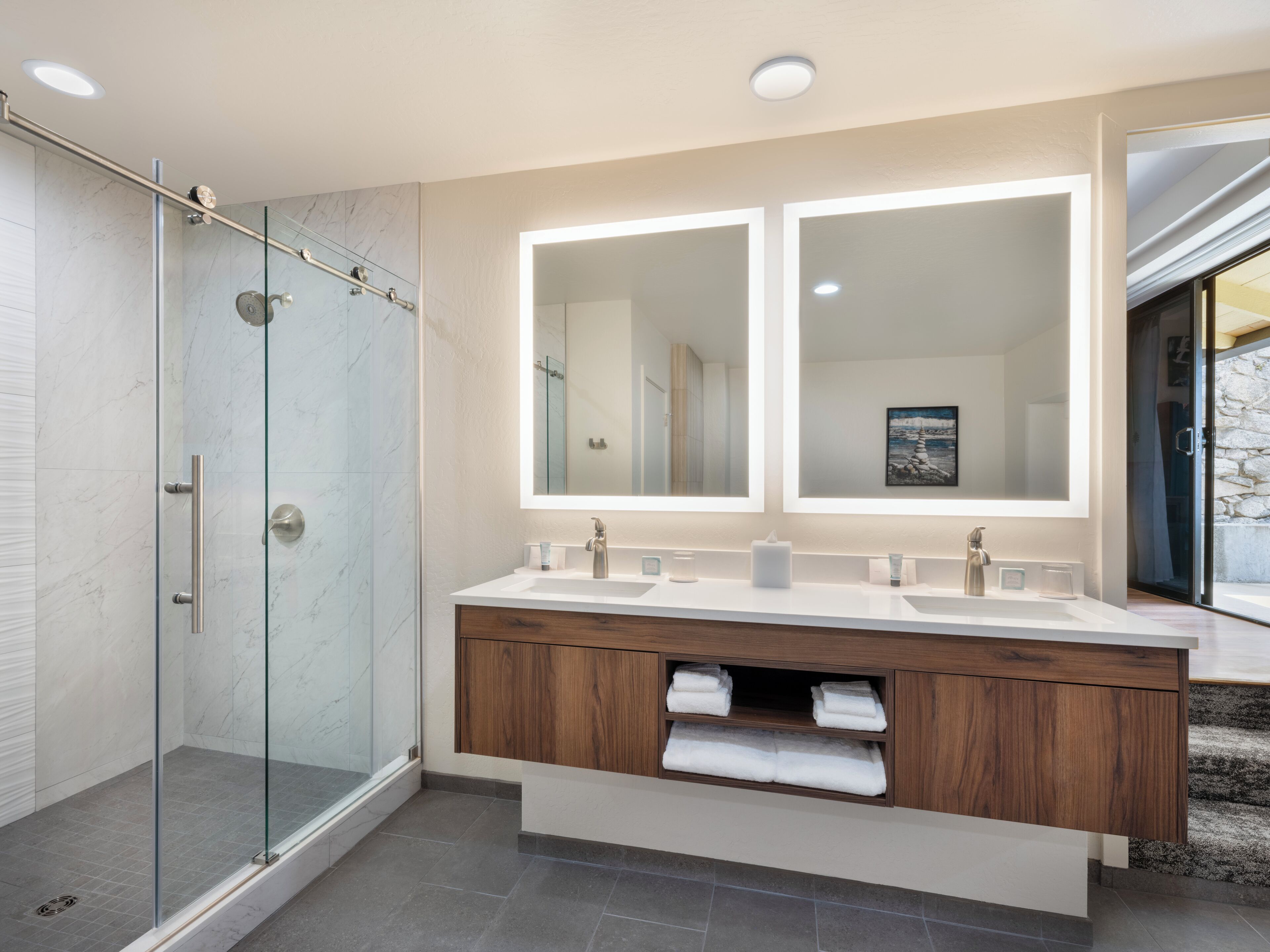 A modern bathroom with a glass shower enclosure, dual sinks with wooden vanity, and a large mirror above.