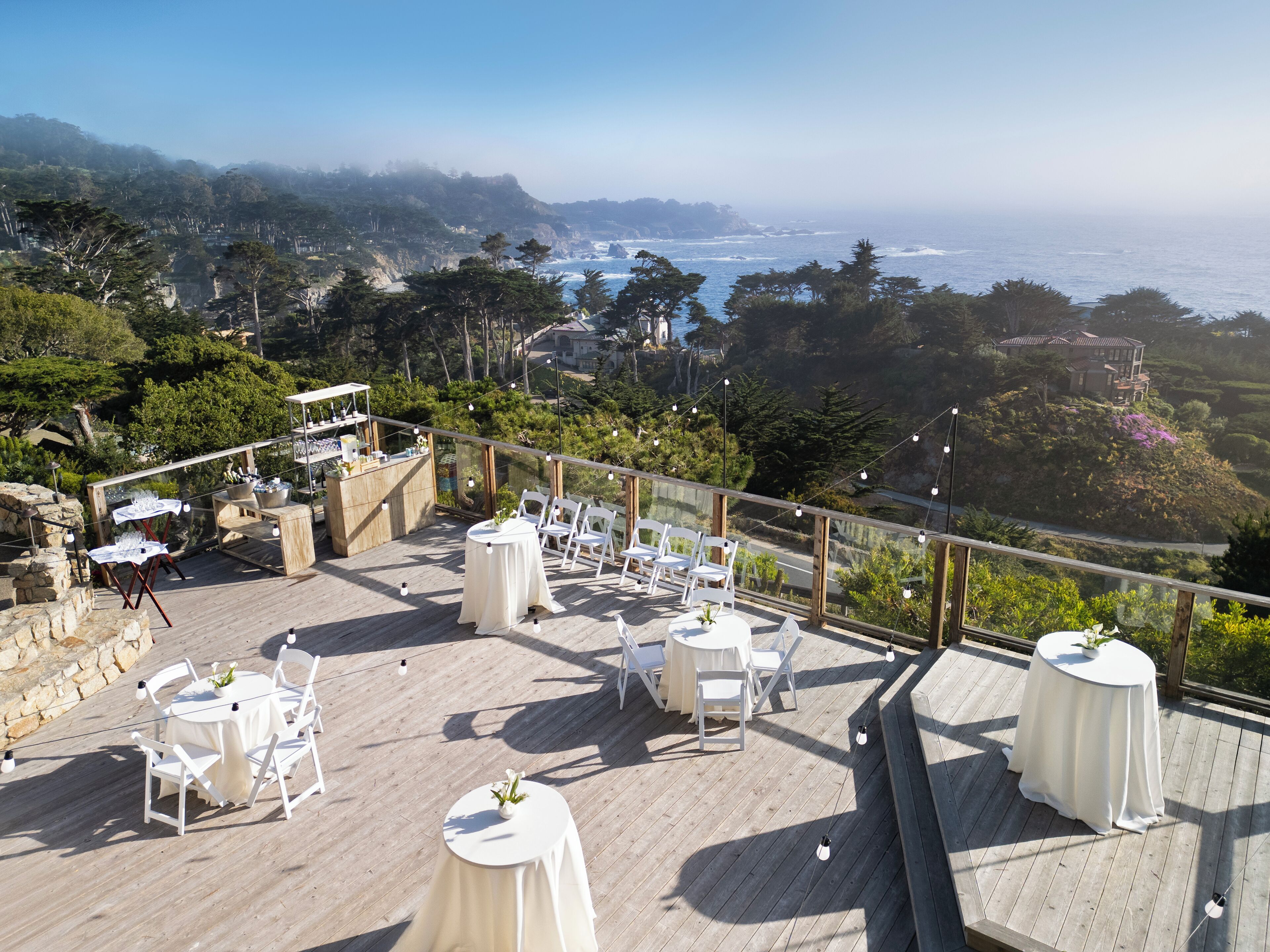 Un patio al aire libre con mesas y sillas, con vistas a un paisaje costero con árboles y el mar.