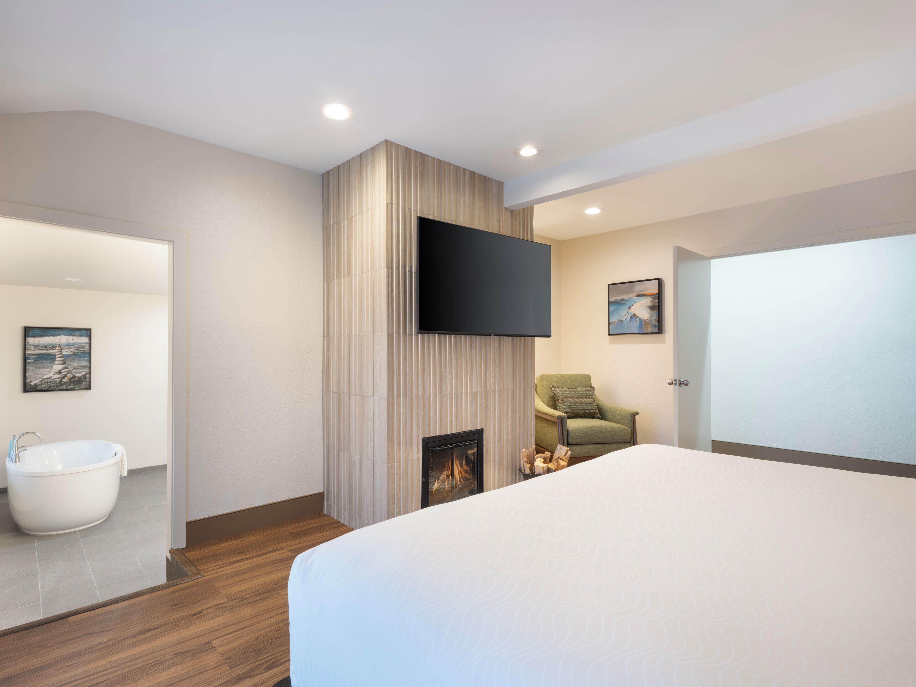 Una habitación de hotel moderna con una cama grande, un televisor de pantalla plana en la pared, una chimenea, un sillón verde y un baño con bañera y lavamanos.