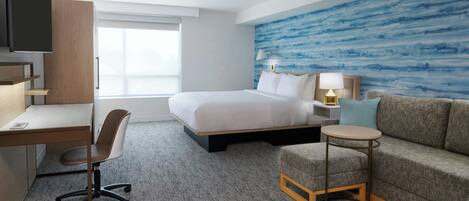 Quarto de hotel com cama, escrivaninha, cadeira, sofá, mesa redonda pequena e televisão fixada na parede.