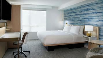 Quarto de hotel com cama, escrivaninha, cadeira e televisão.