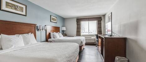 A hotel room with two beds, a desk, a chair, a lamp, and a window with a view.