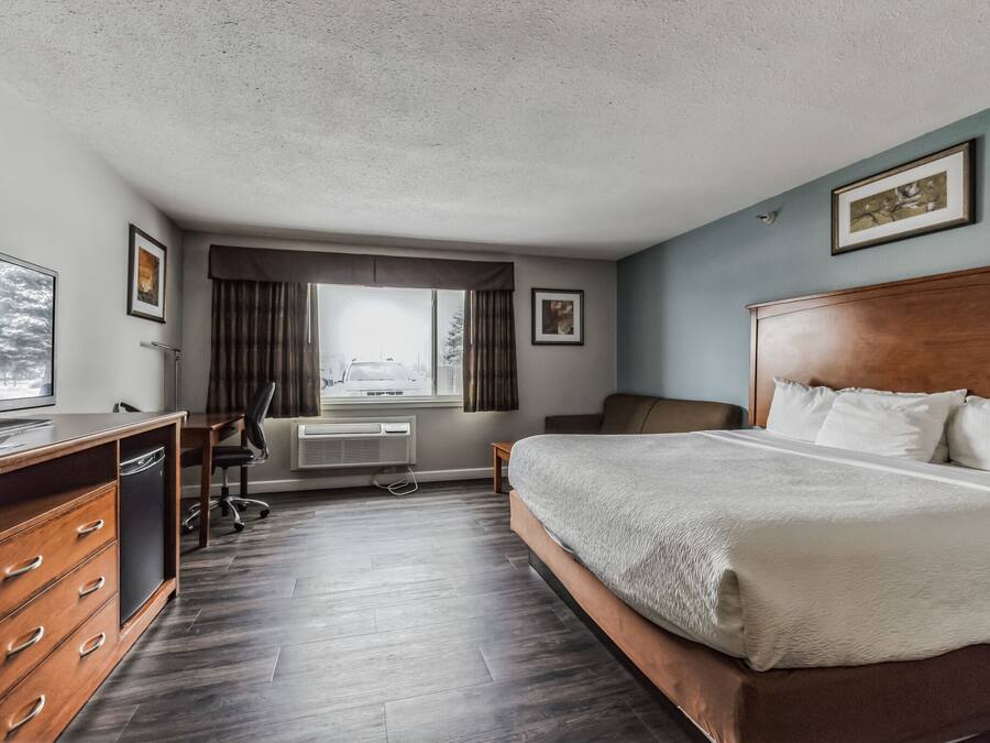 Ett hotellrum med en säng i trä, ett skrivbord med en stol, en tv och ett fönster med gardiner.