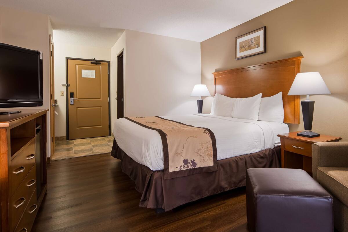 A hotel room with a bed, two bedside lamps, a TV, a wooden desk, a chair, and a small ottoman.