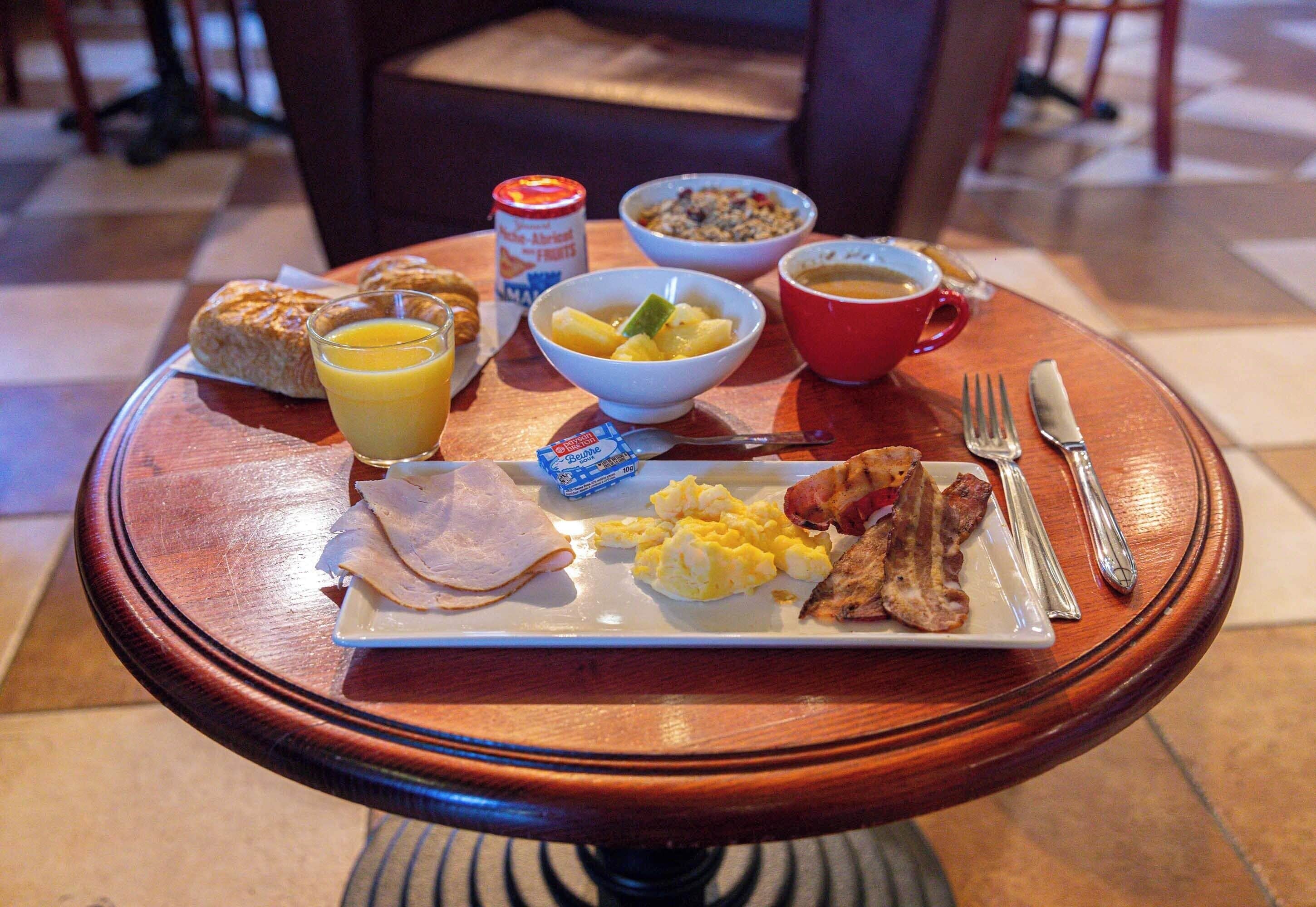 A breakfast tray with croissants, orange juice, scrambled eggs, bacon, and a bowl of fruit on a wooden table.