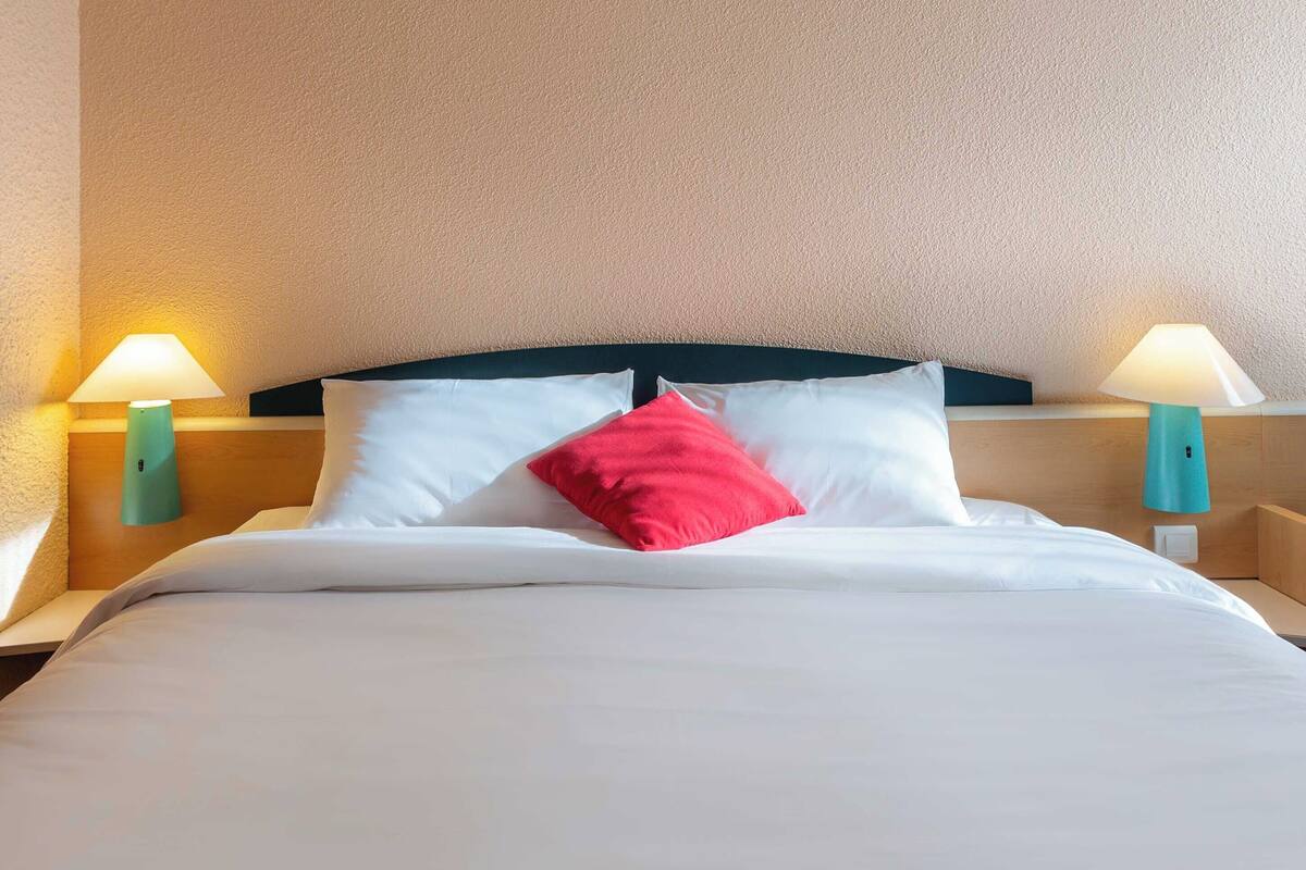 Un lit bien fait, avec du linge de lit blanc et un seul oreiller rouge, flanqué de deux lampes de chevet turquoise posées sur des tables de nuit en bois.
