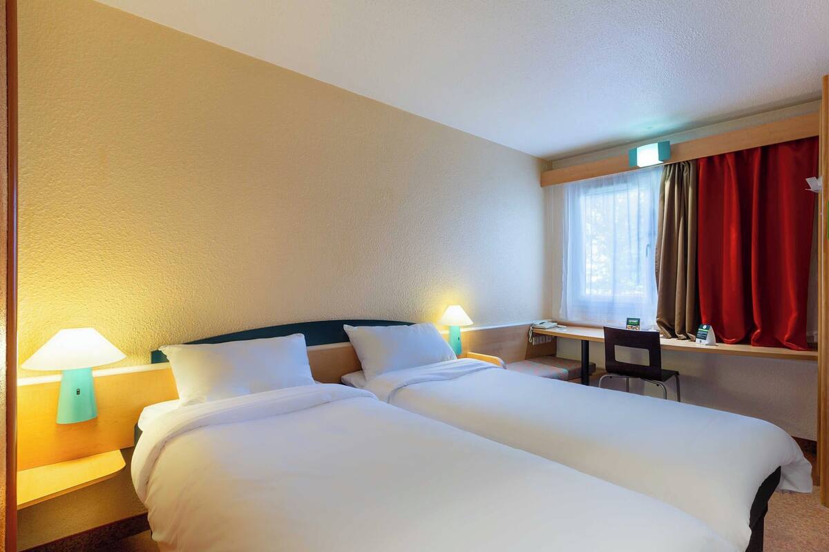 Une chambre d’hôtel avec deux lits, un bureau, une chaise et une fenêtre avec des rideaux.