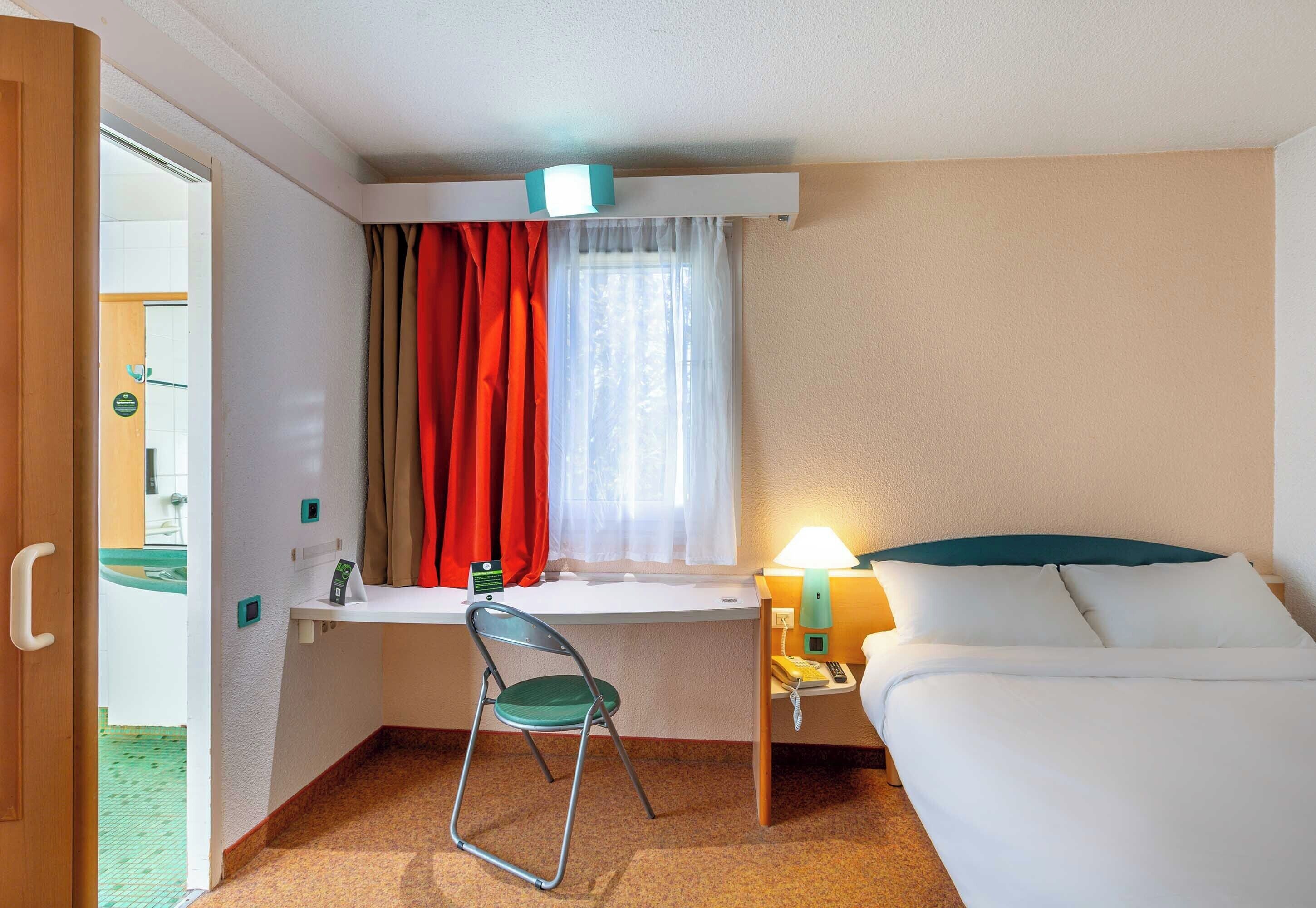 Una camera d'albergo compatta con un letto, una scrivania con sedia, una finestra con tende, una lampada da comodino e una porta che dà su un'altra stanza.