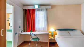 Una camera d'albergo compatta con un letto, una scrivania con sedia, una finestra con tende, una lampada da comodino e una porta che dà su un'altra stanza.