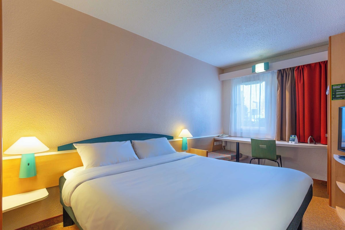 Une chambre d’hôtel avec un grand lit, deux lampes de chevet, un bureau avec une chaise et une fenêtre avec des rideaux.