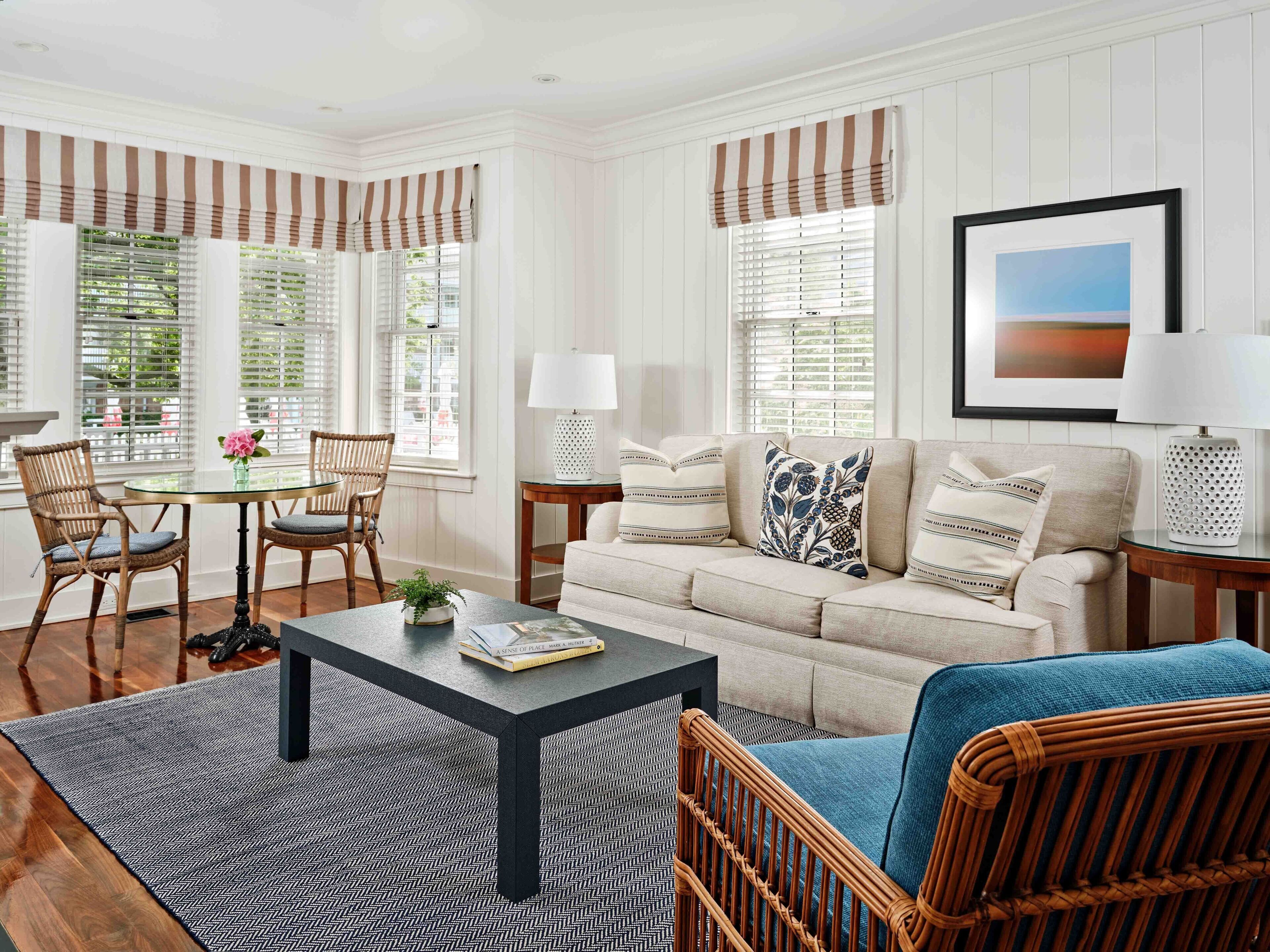 A living room with a sofa, two armchairs, a coffee table, and a dining table with chairs. There are windows with blinds, a lamp, and a framed picture on the wall.