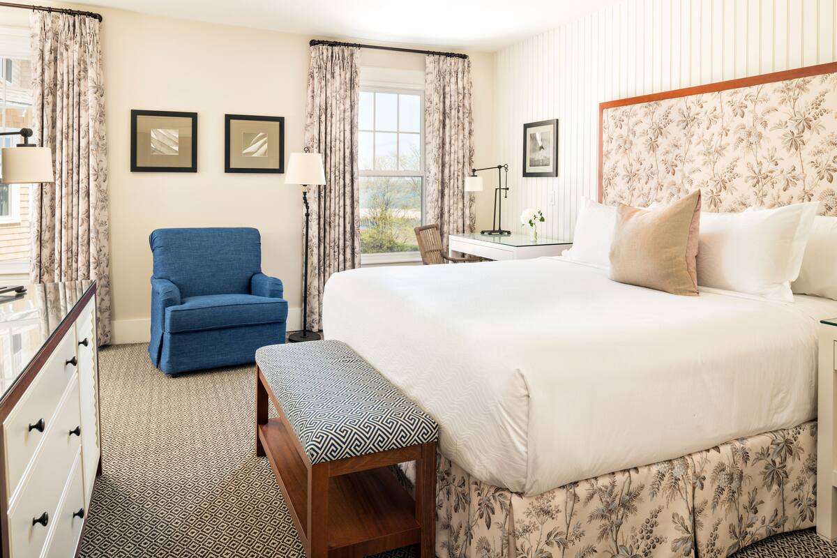 A hotel room with a large bed, a blue armchair, a wooden bench, a desk with a lamp, and a window with curtains.