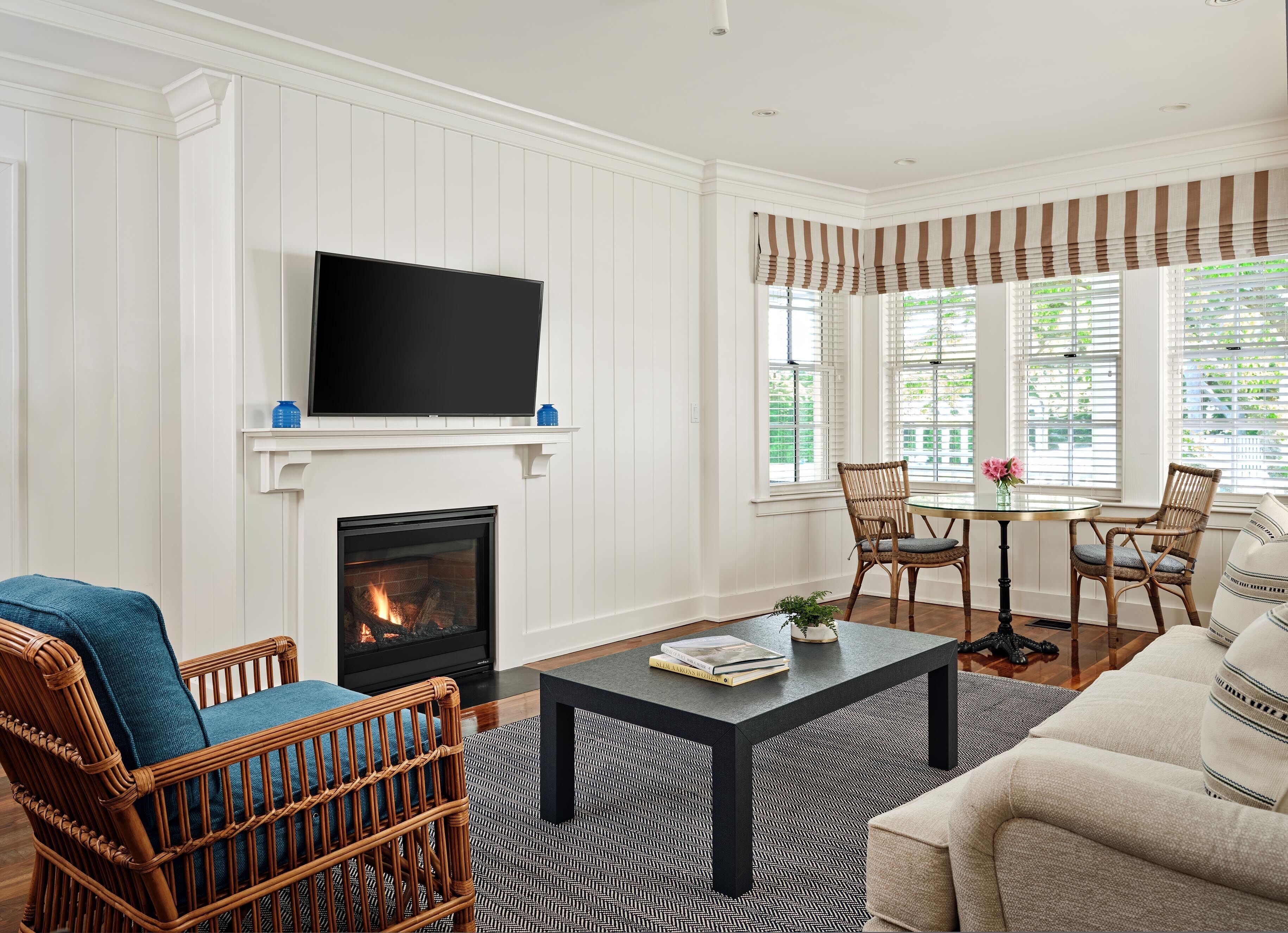 A living room with a fireplace, a television mounted above it, wicker chairs, a round table with chairs, and large windows with blinds.