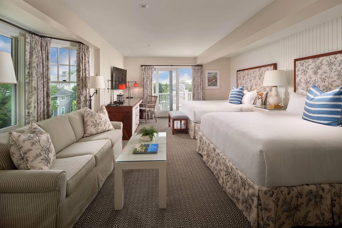 A hotel room with two beds, a sofa, a small table, and a desk with a chair. There are curtains on the windows, a lamp on the desk, and a plant on the table.