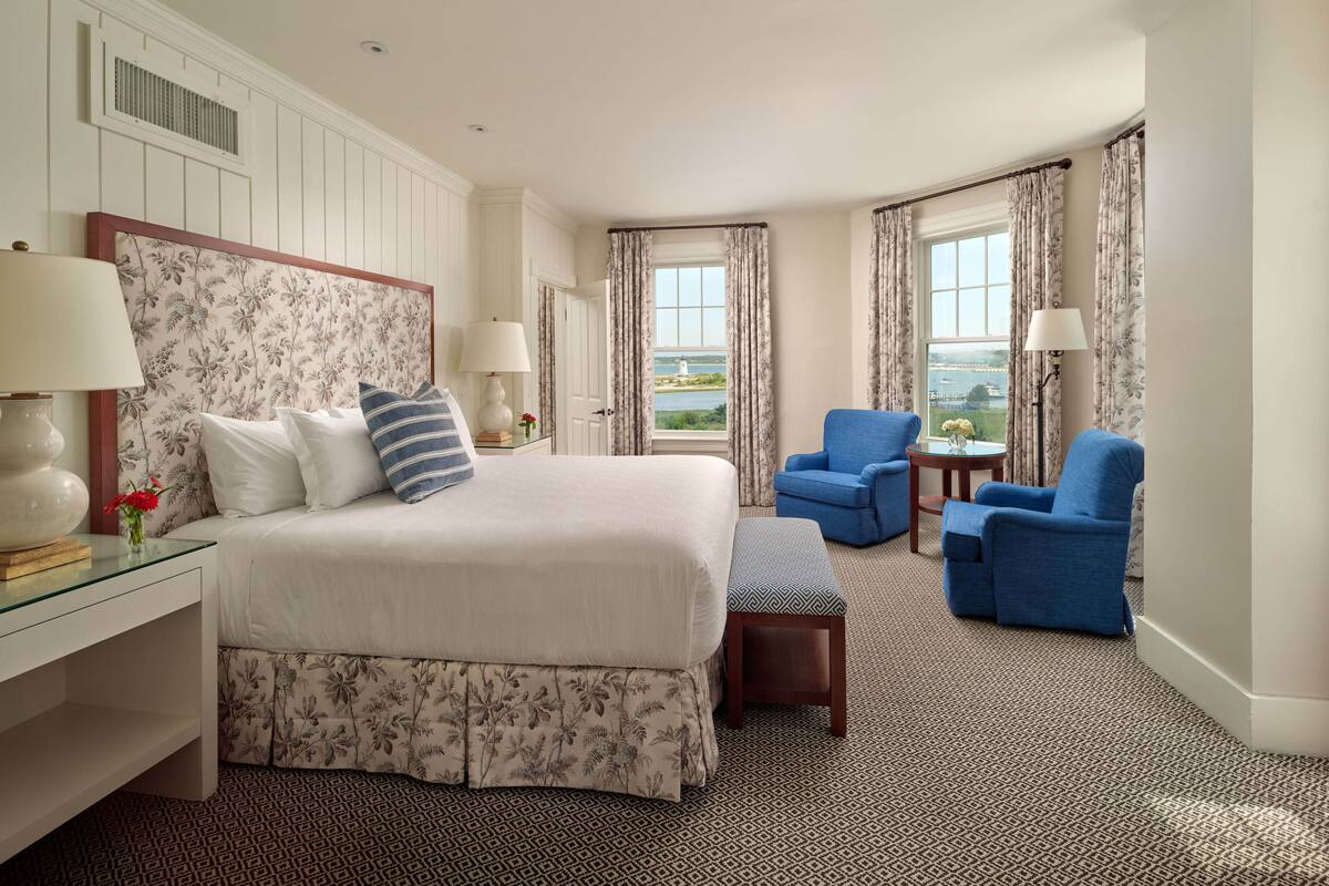 A hotel room with a large bed, two blue armchairs, a small table, and a view of the outdoors.