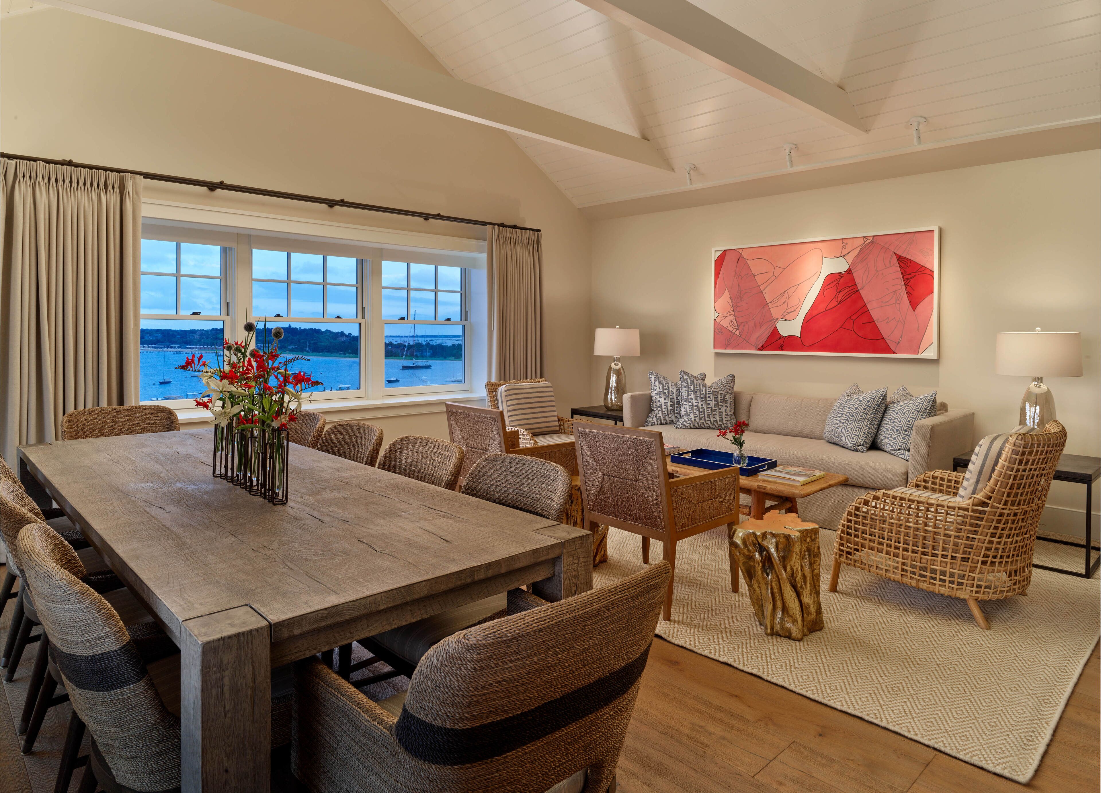 A spacious living room with a large window overlooking a body of water, a dining area with a wooden table and wicker chairs, and a cozy seating area with a sofa and patterned cushions.