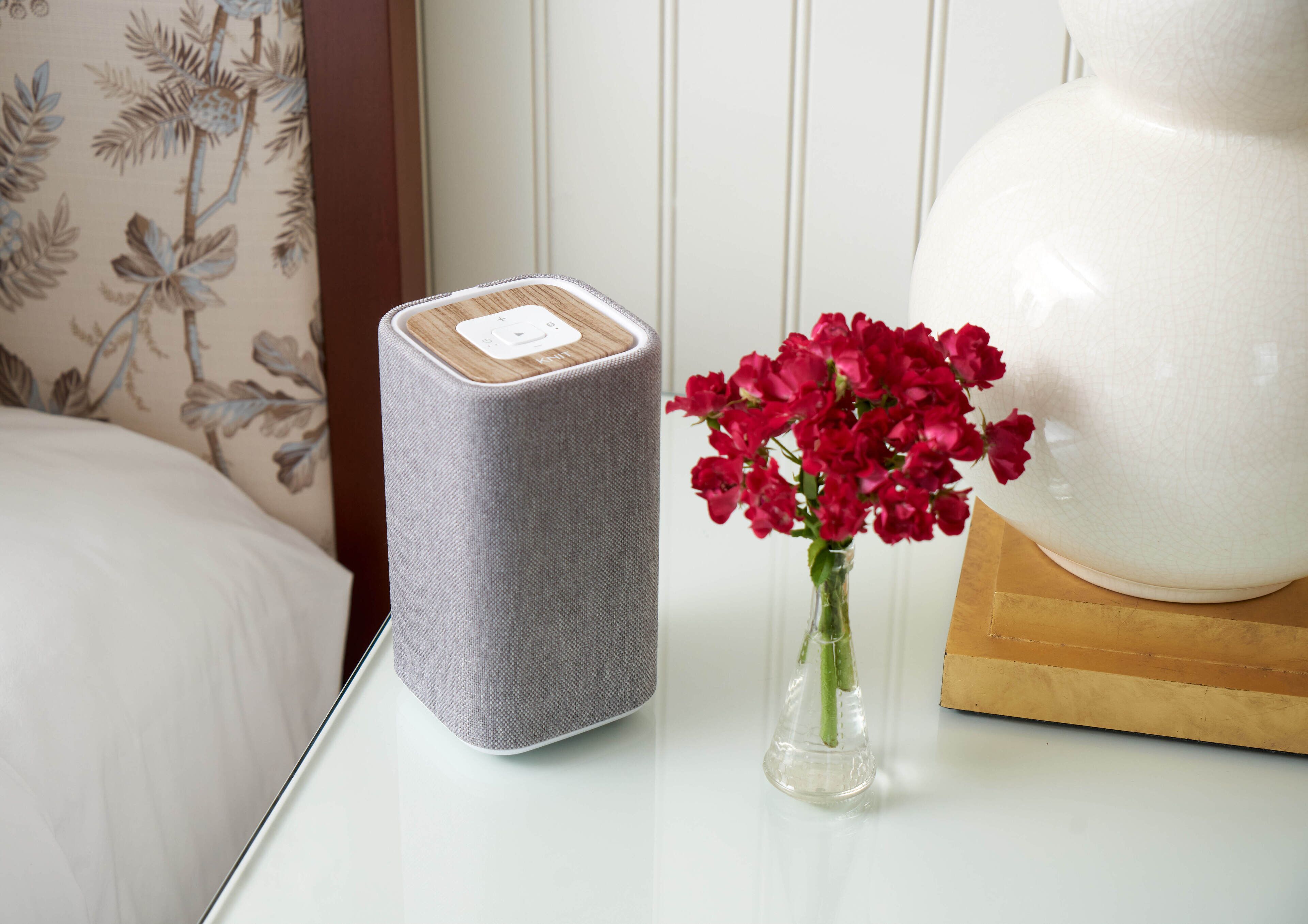 Une table blanche avec un haut-parleur gris, un vase transparent contenant des fleurs rouges et une lampe blanche.