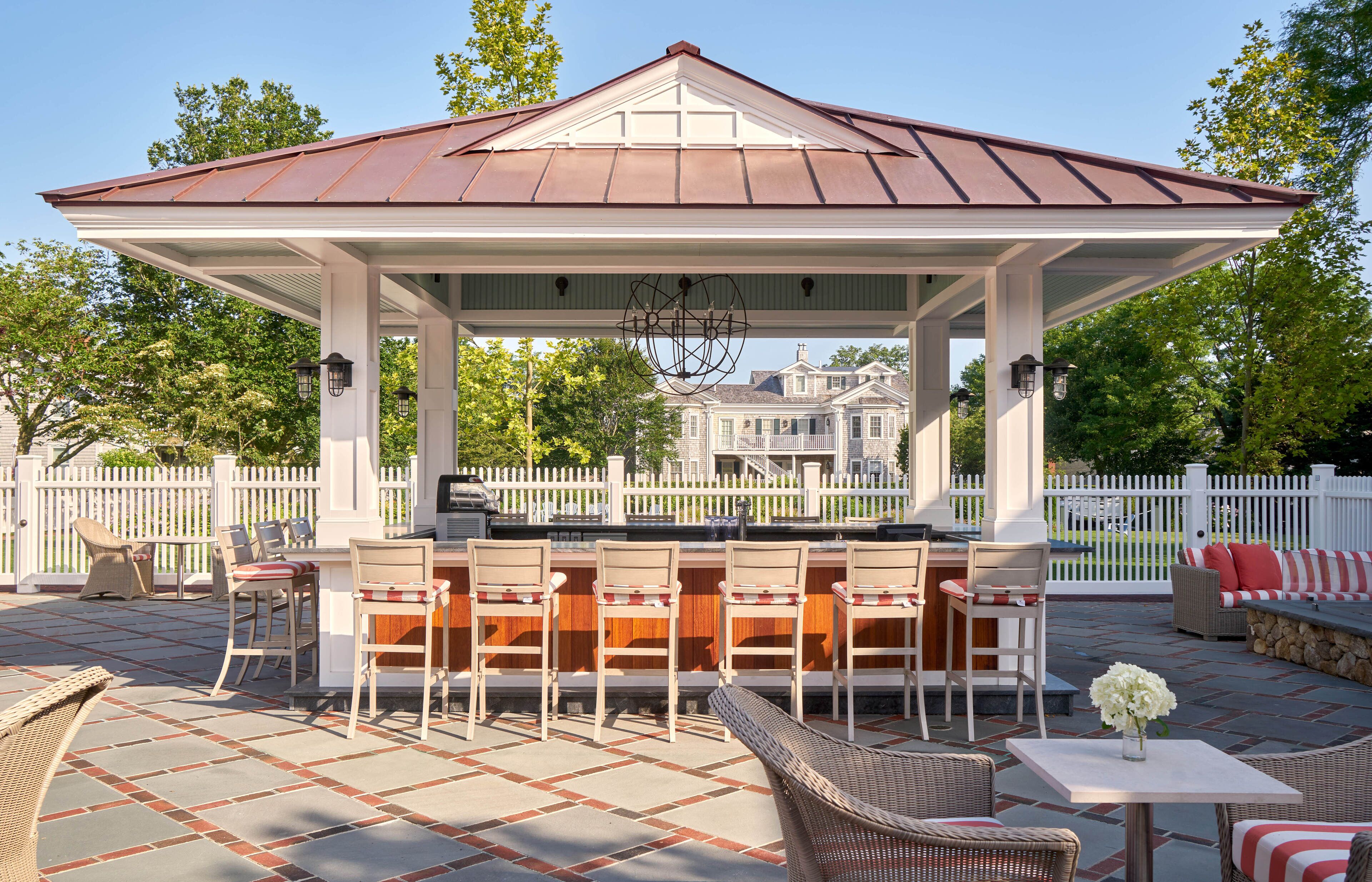 A covered outdoor patio with a bar area, seating, and a view of a large house in the background.