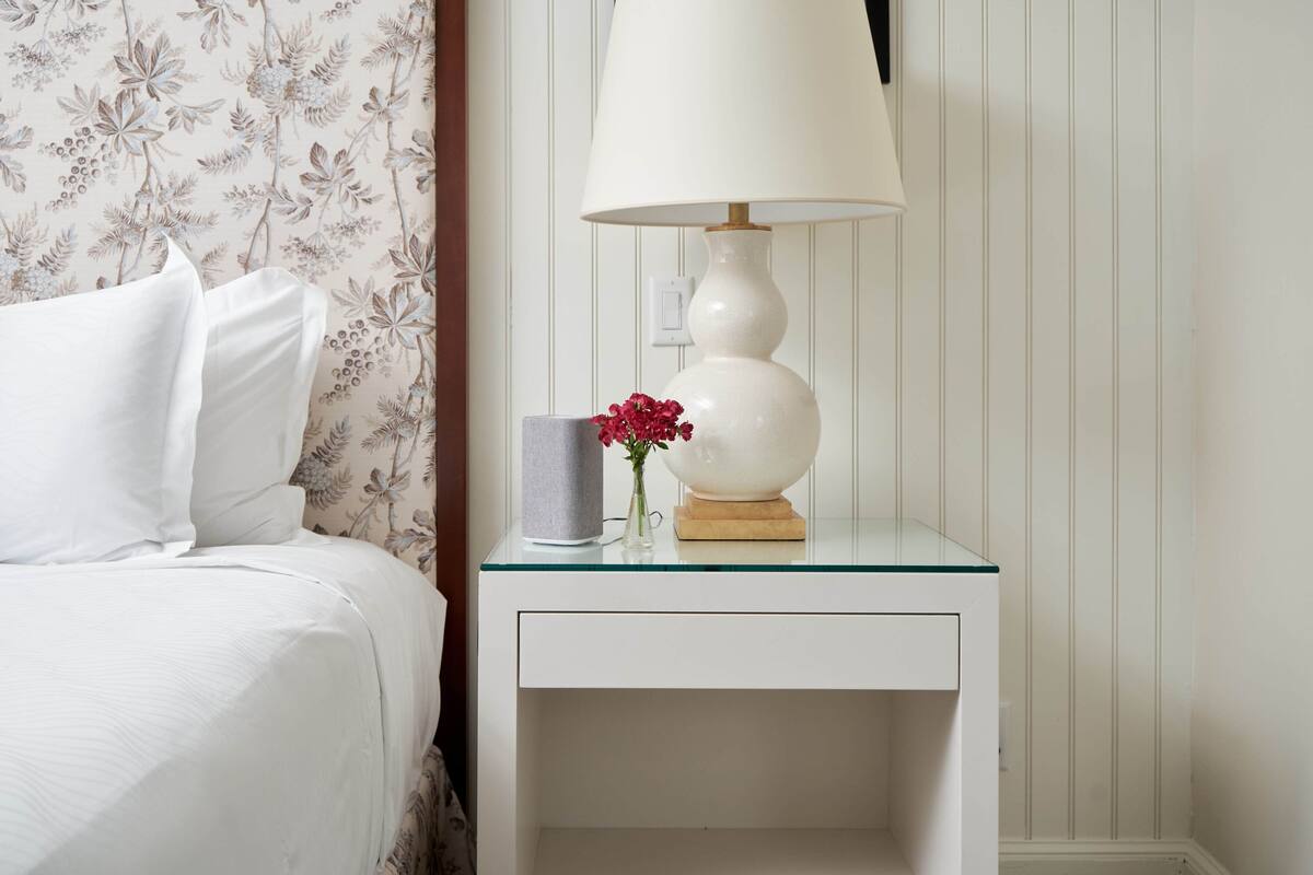 A neatly made bed with white linens, a white nightstand with a lamp and a vase of flowers, and a floral wallpaper pattern.