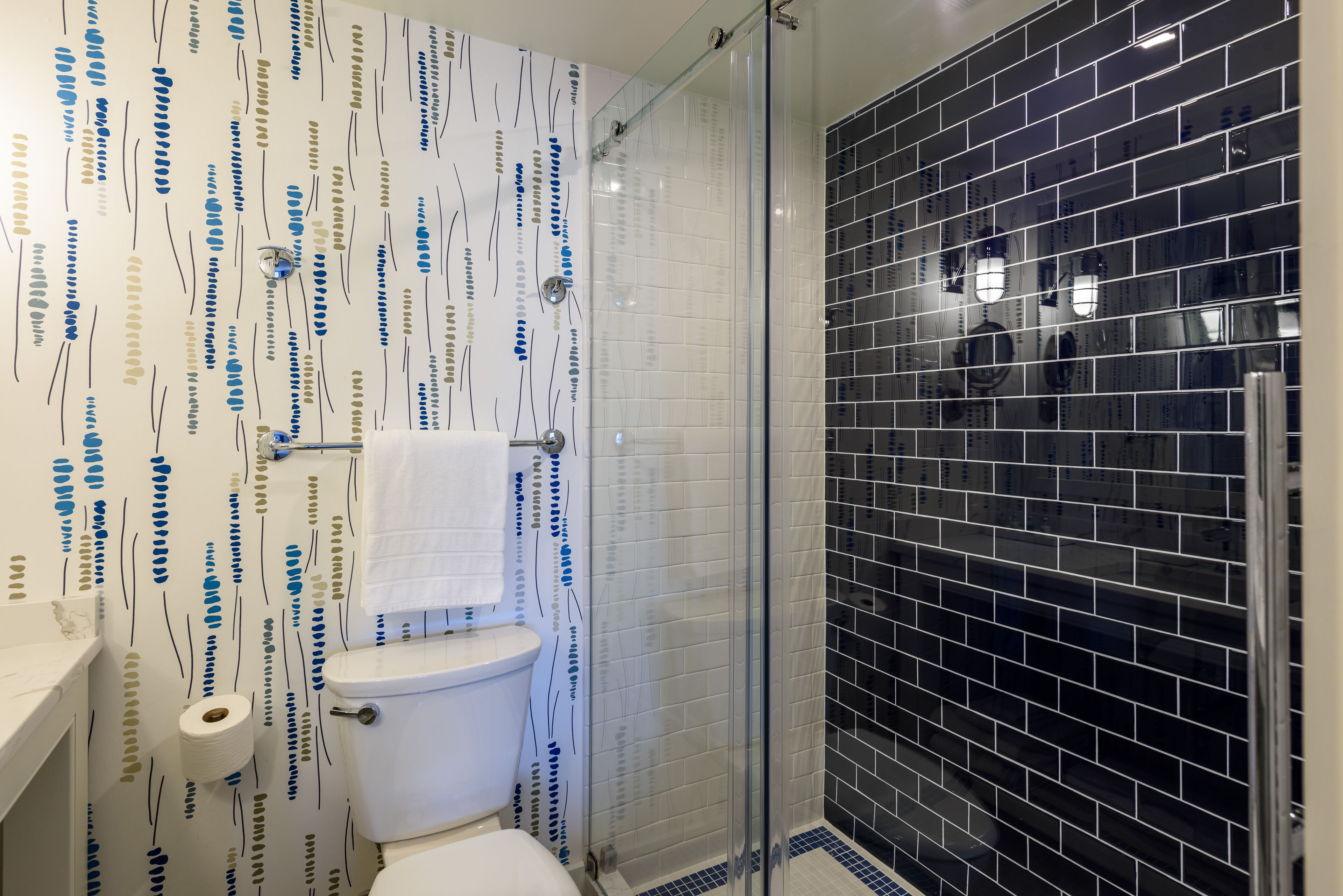 A bathroom with a patterned wall, a toilet, a shower with a glass door, and a sink with a towel hanging on the wall.