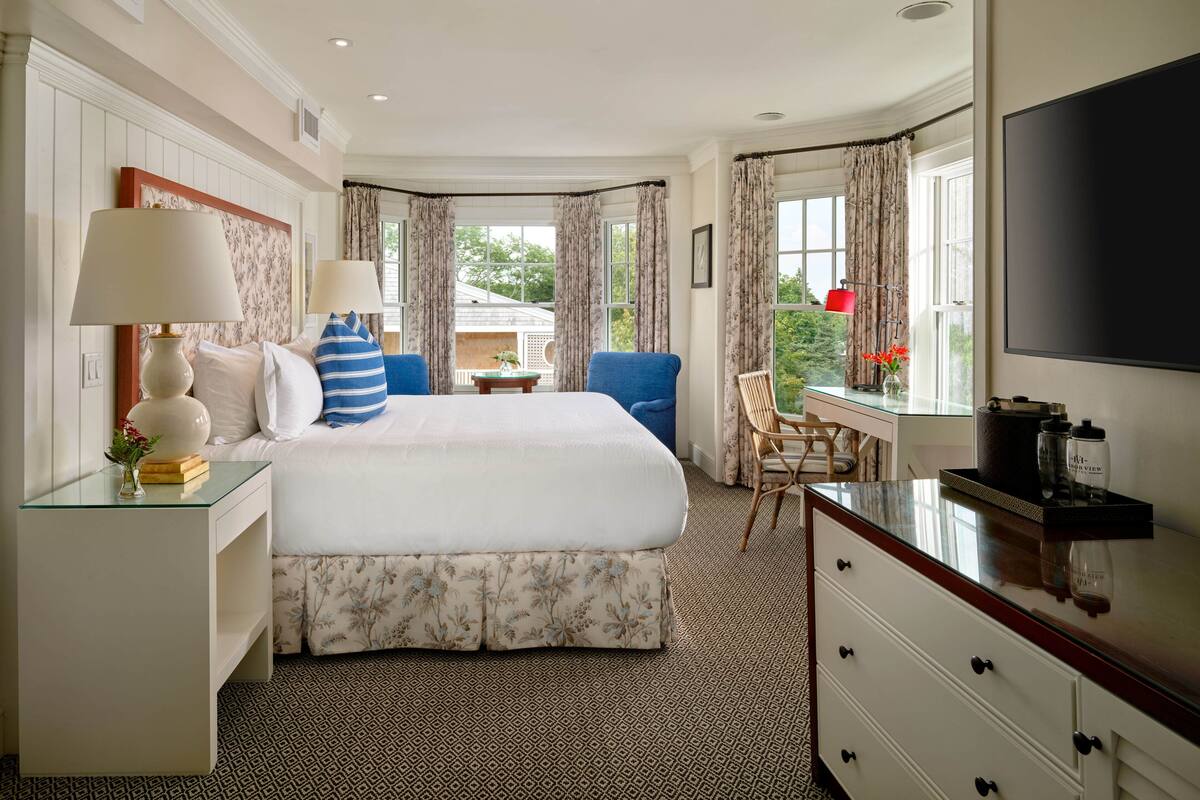 A hotel room with a large bed, two bedside tables with lamps, a desk with a chair, a TV, and a window with curtains.