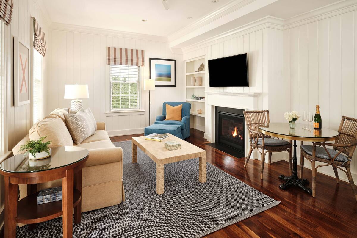 A living room with a sofa, a coffee table, a fireplace, a TV mounted above the fireplace, and a dining area with chairs and a table.