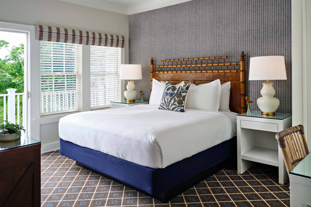 A hotel room with a large bed, two bedside tables with lamps, a wooden headboard, a window with blinds, and a patterned carpet.