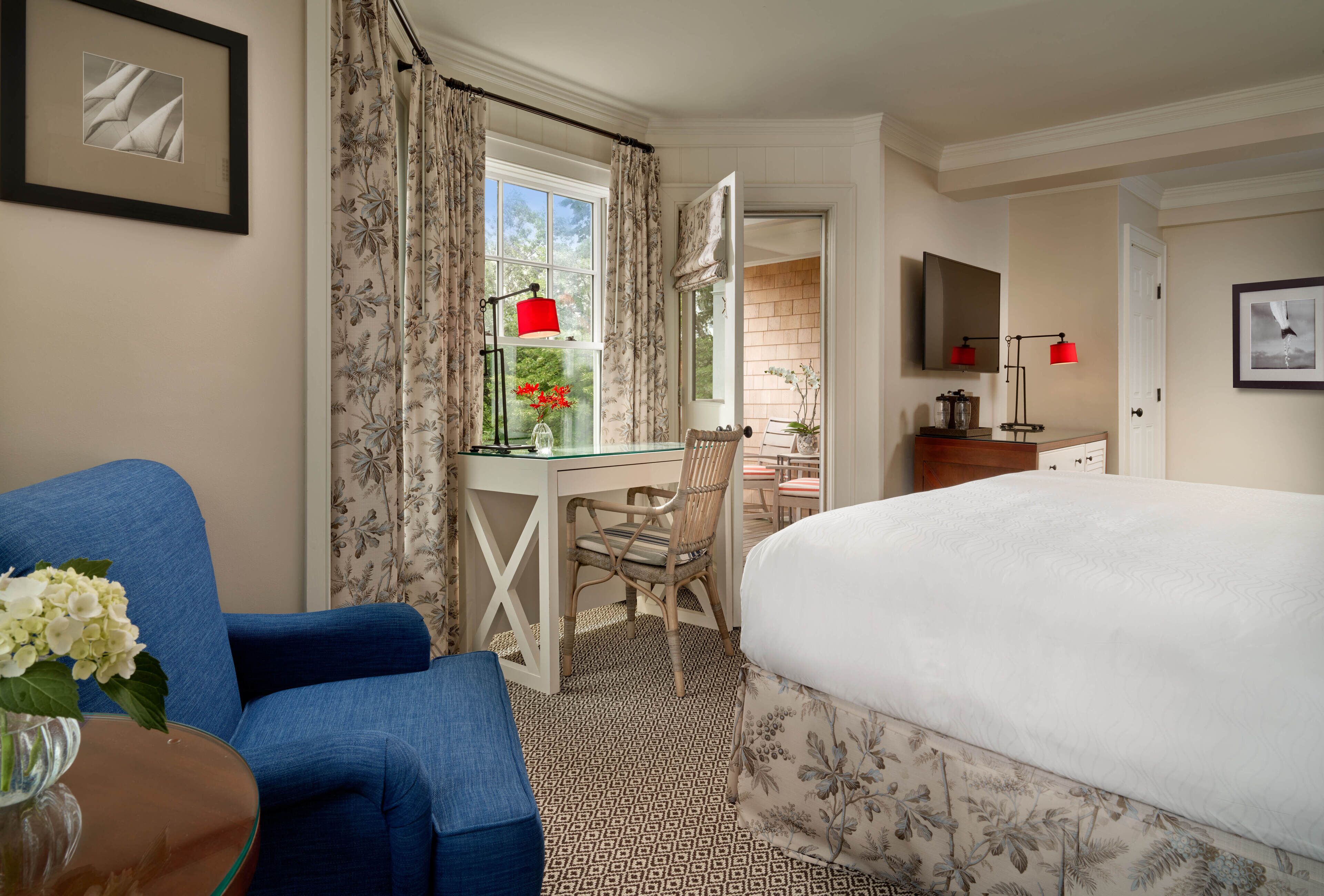 A hotel room with a bed, a desk with a chair, a blue armchair, a small table with a vase of flowers, and a window with curtains.