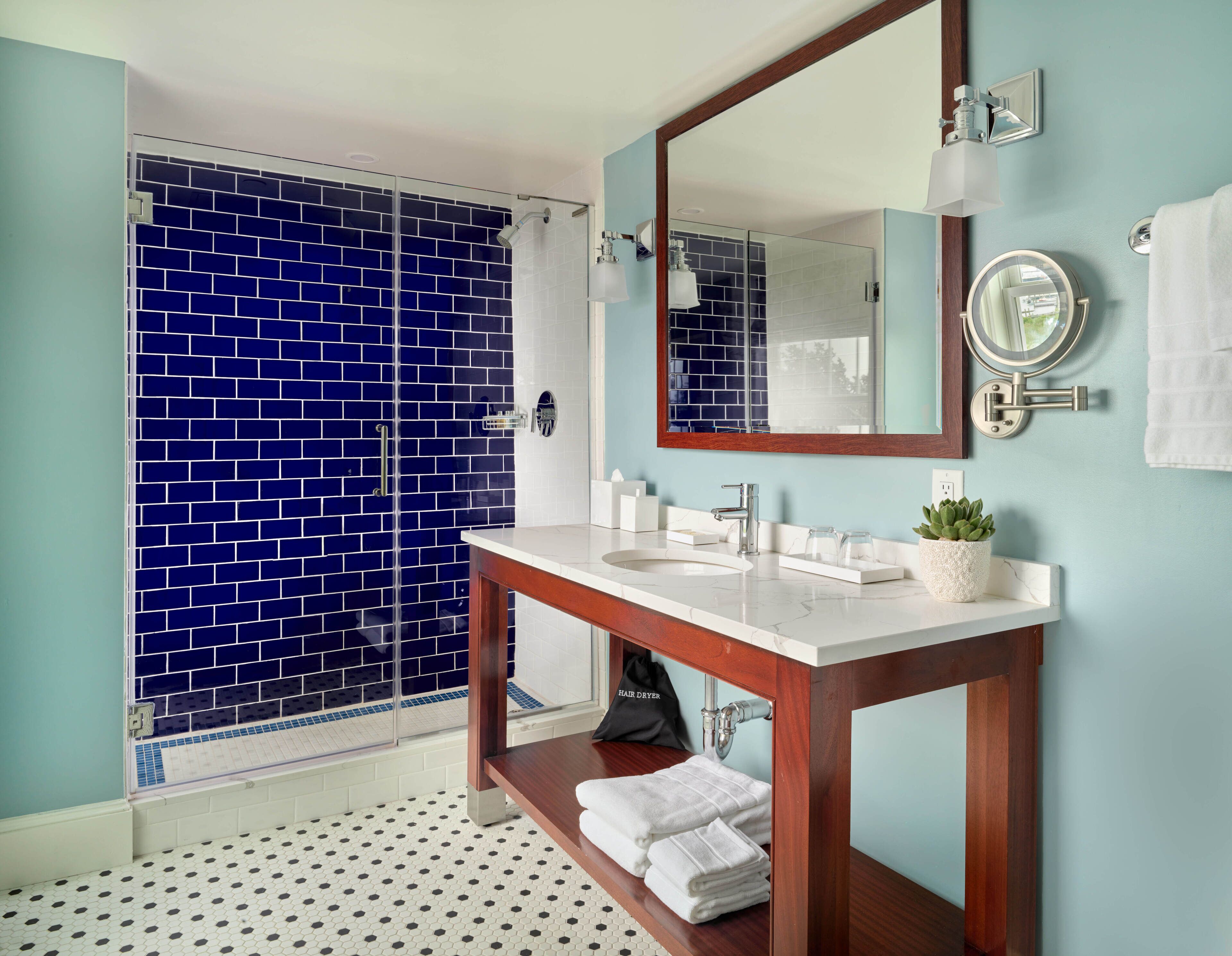 A bathroom with a blue-tiled shower, a white sink with a wooden vanity, a mirror, and a potted plant.