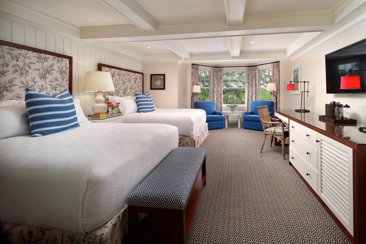 A hotel room with two beds, a bench, a desk with drawers, a chair, a small table, and a TV mounted on the wall.
