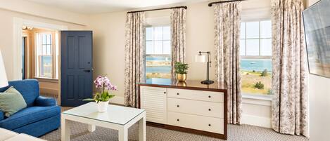 A room with a blue sofa, a white coffee table, a white dresser, and a view of the sea through large windows with floral curtains.