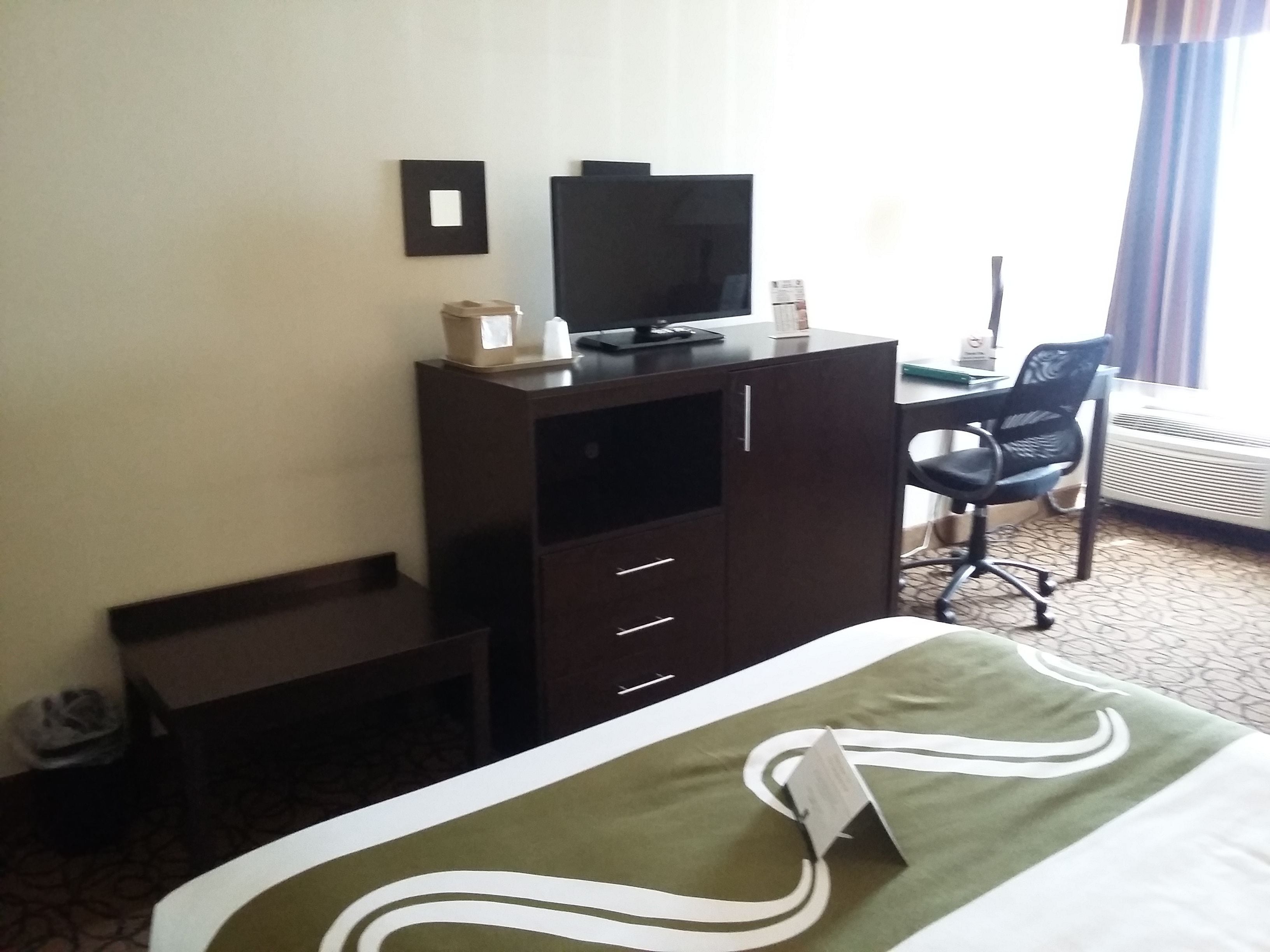 Habitación de hotel con cama, escritorio, silla, televisión y ventana con cortinas.