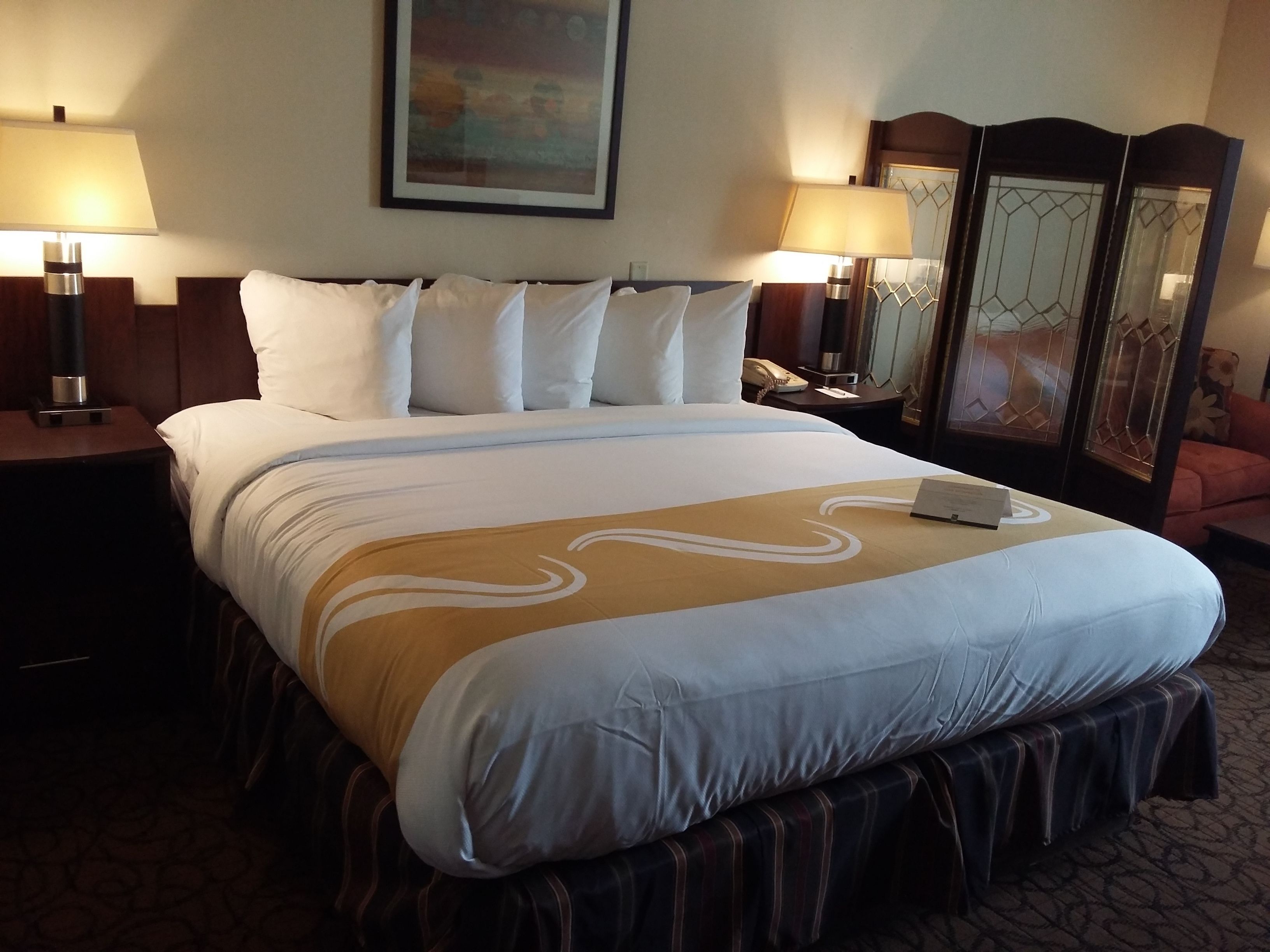 A hotel room with a large bed, two bedside tables, a nightstand, a lamp, a mirror, and a painting on the wall.