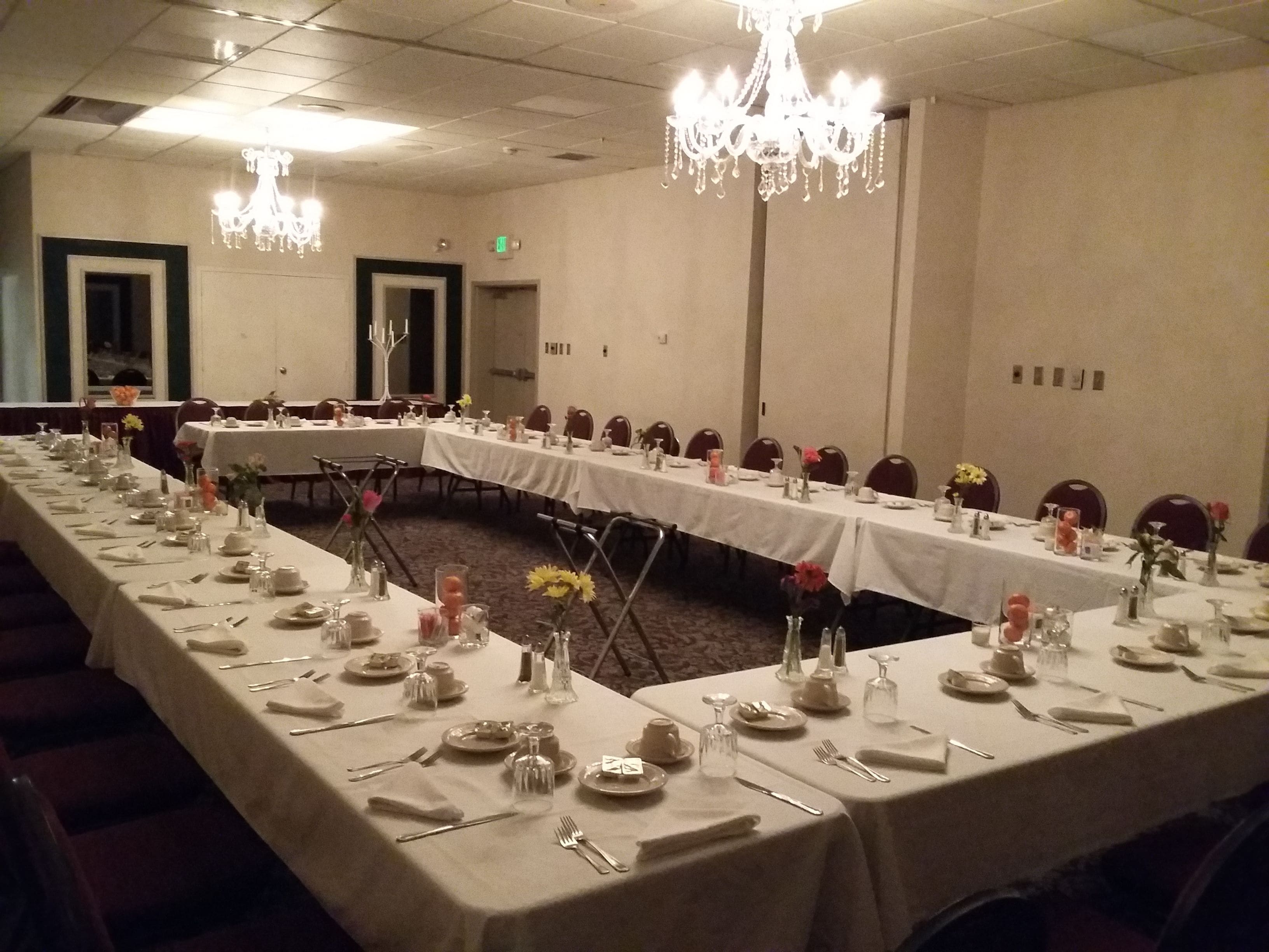 Uma sala de banquetes com mesas compridas preparadas para refeições, lustres e piso coberto de carpete.