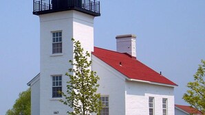 A white lighthouse with a red roof and a black top, surrounded by a white fence and a small tree.