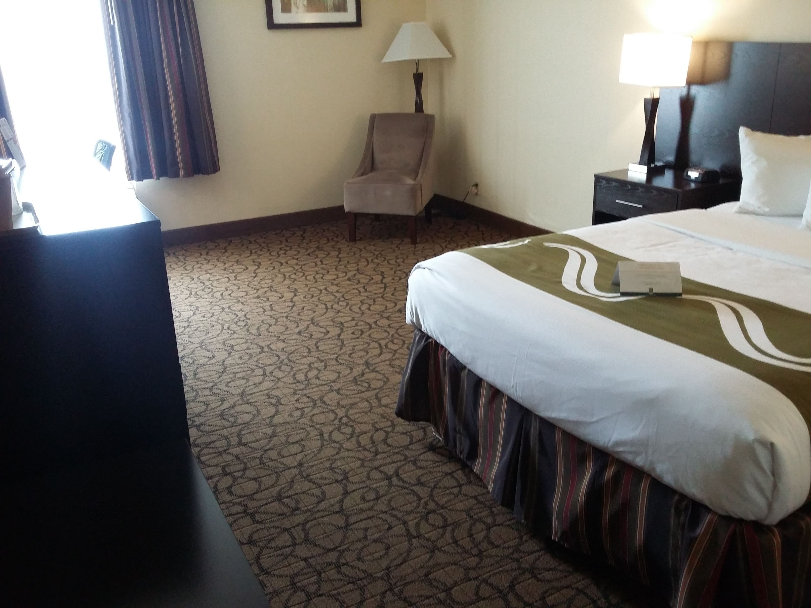 A hotel room with a bed, a chair, a desk, a lamp, and a nightstand.